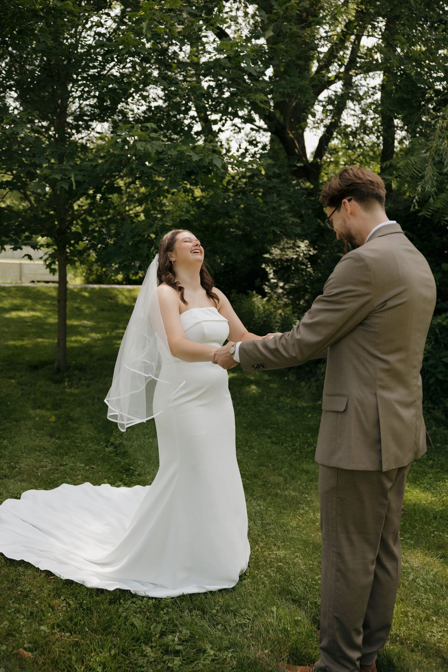 Joyful bride in white gown and veil laughing while holding hands with groom in tan suit outdoors.
