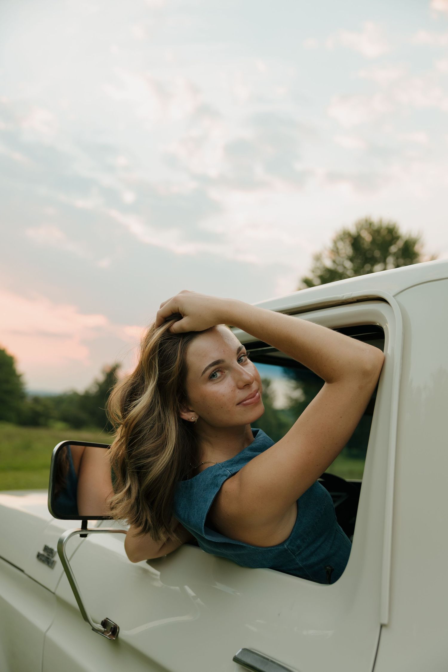 Young woman with long hair leans out of a white vintage truck window, smiling at sunset.