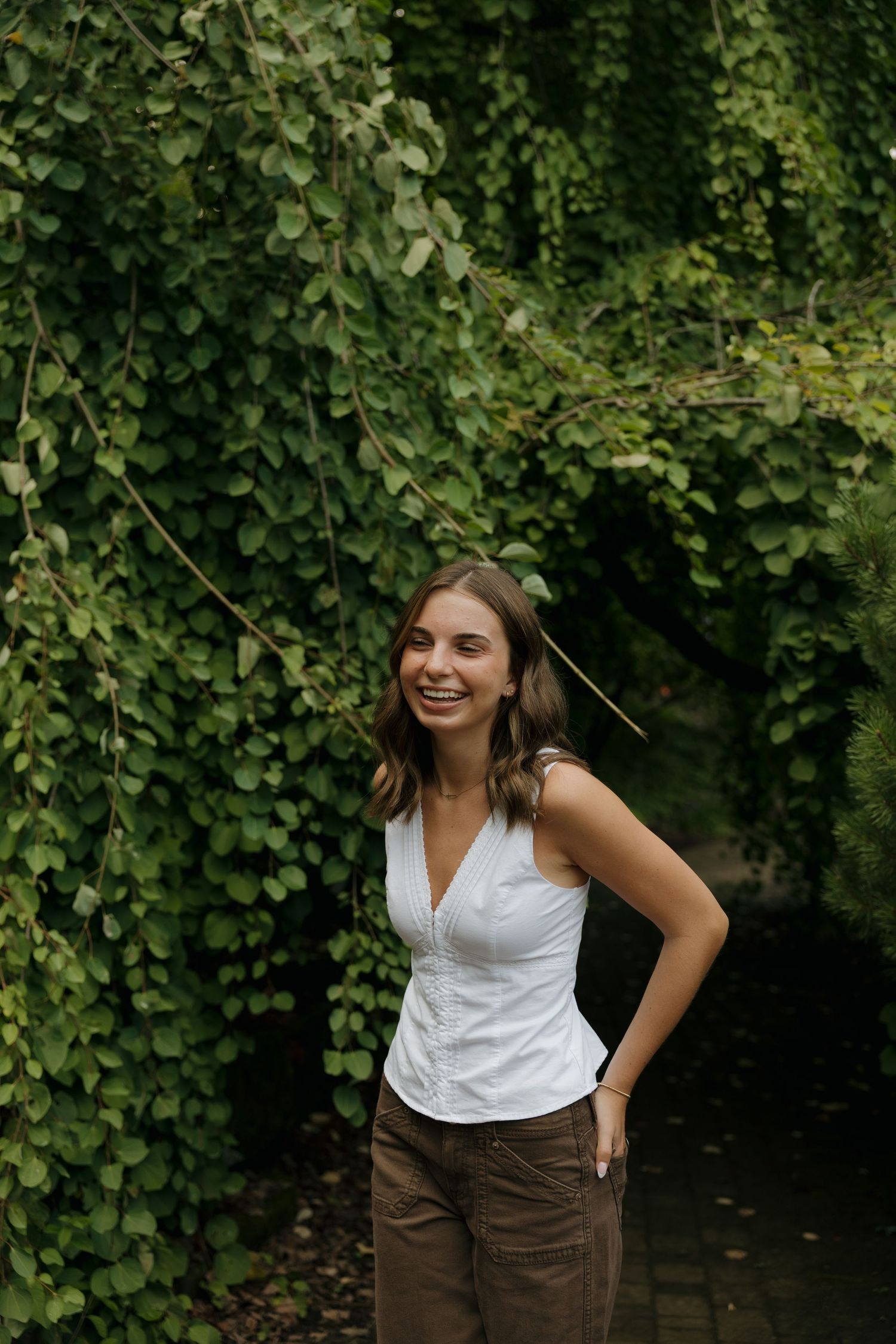 Young woman smiling in white sleeveless top and brown pants standing before lush green ivy wall outdoors.