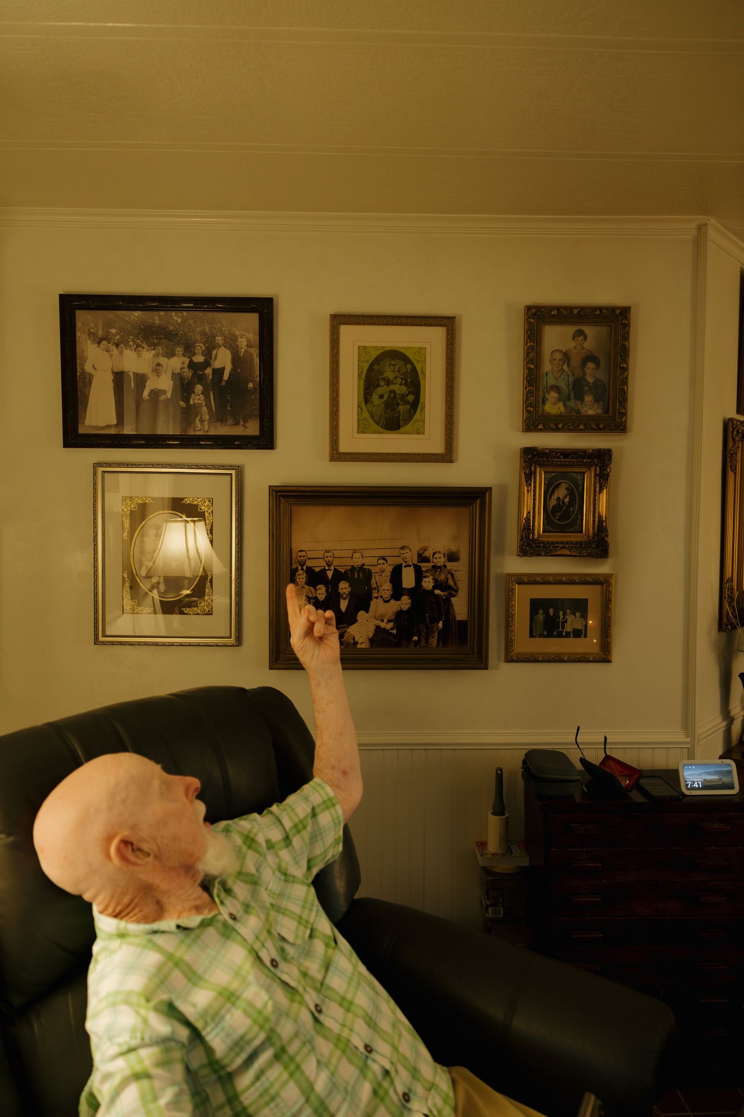 Elderly bald man in green plaid shirt pointing at vintage family photos displayed on a living room wall.