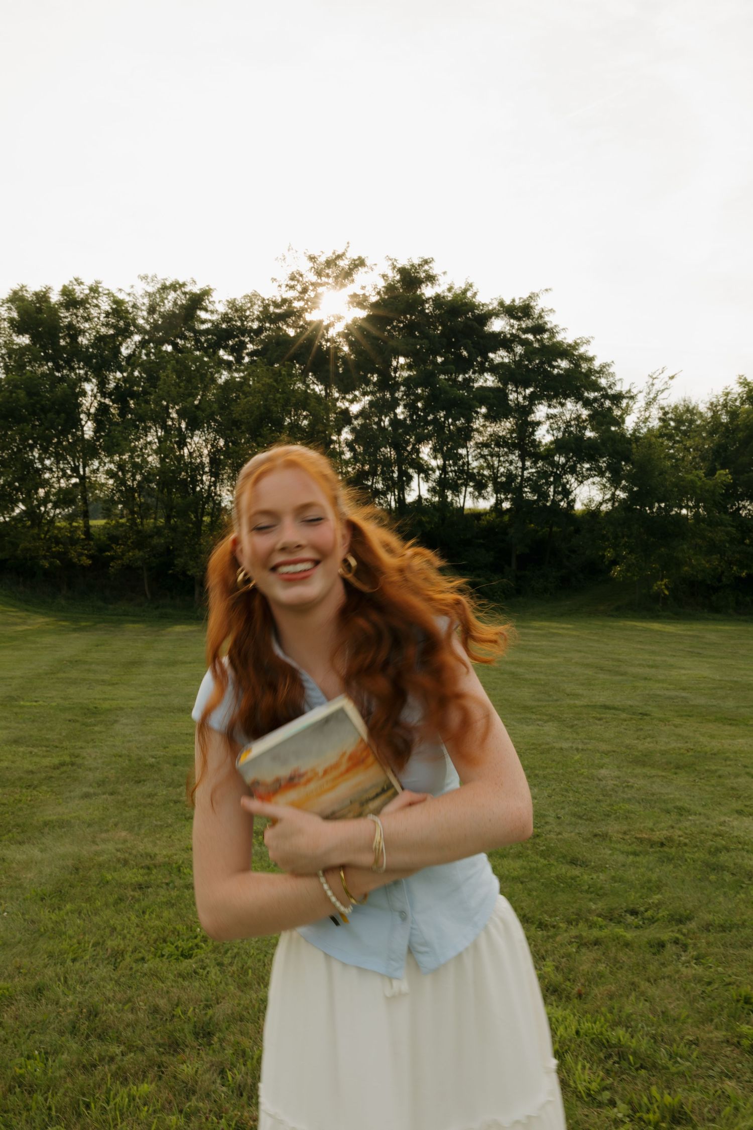 Smiling redhead woman hugging a book outdoors on green grass with trees and golden sunset light behind her.