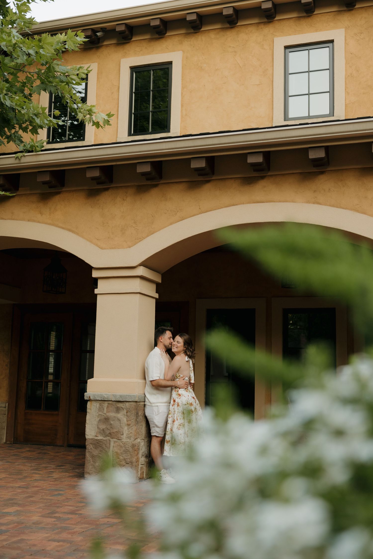 Couple sharing a romantic kiss under arched portico of elegant yellow stucco building surrounded by lush greenery.