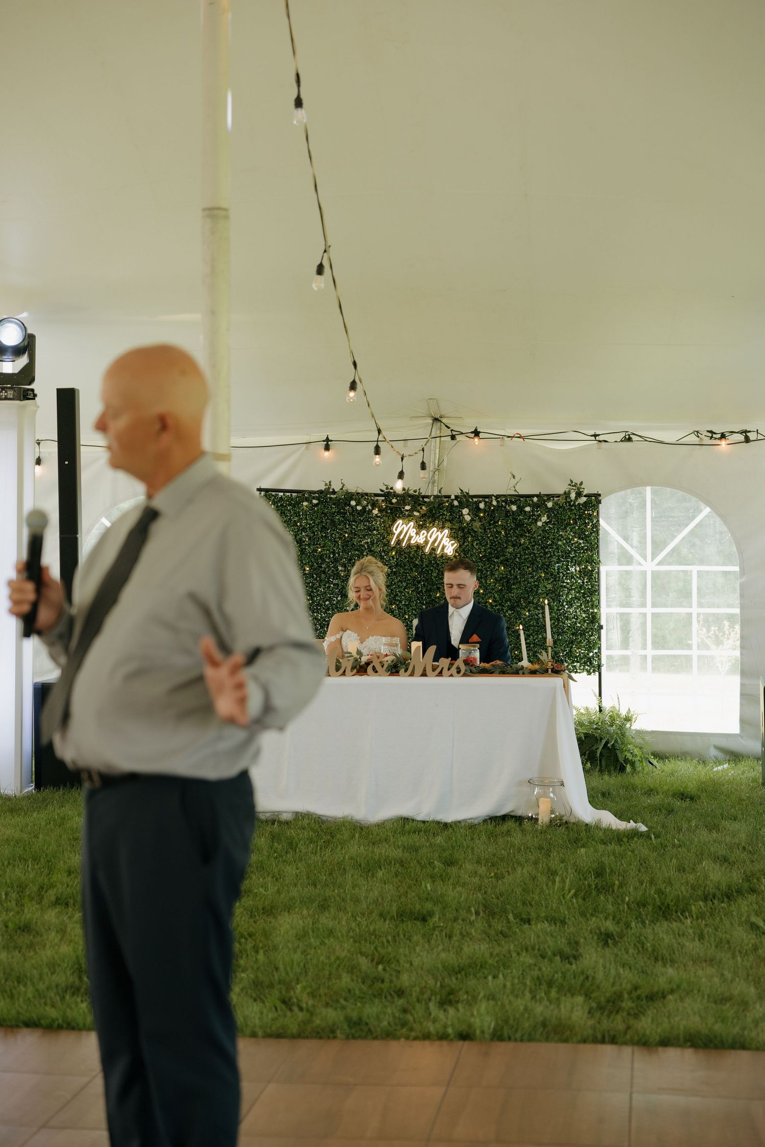 Father giving a toast speech at wedding reception while couple sits at sweetheart table.