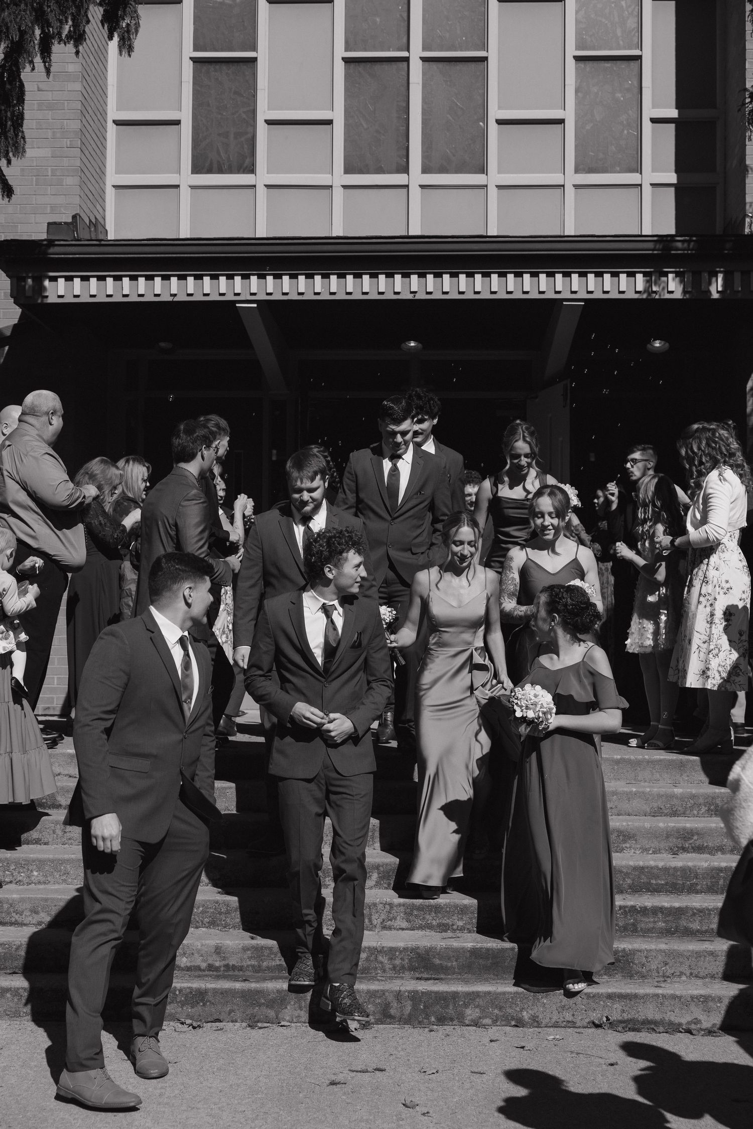 Wedding party in formal attire walks down brick building steps, with the bridesmaids holding bouquets, in black and white.