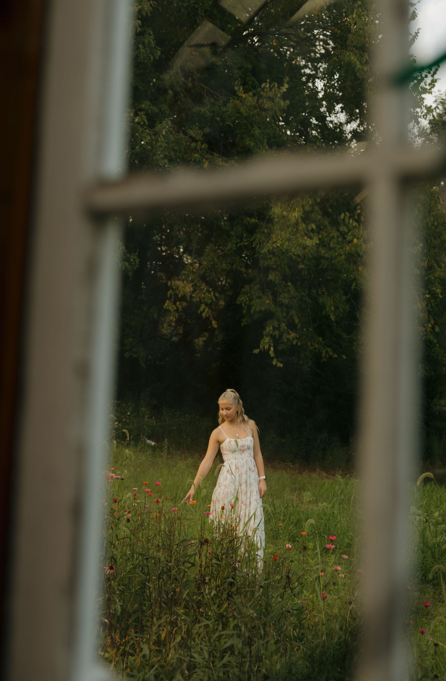 A woman in a white floral dress walks through a wildflower meadow, viewed through a rustic window frame.