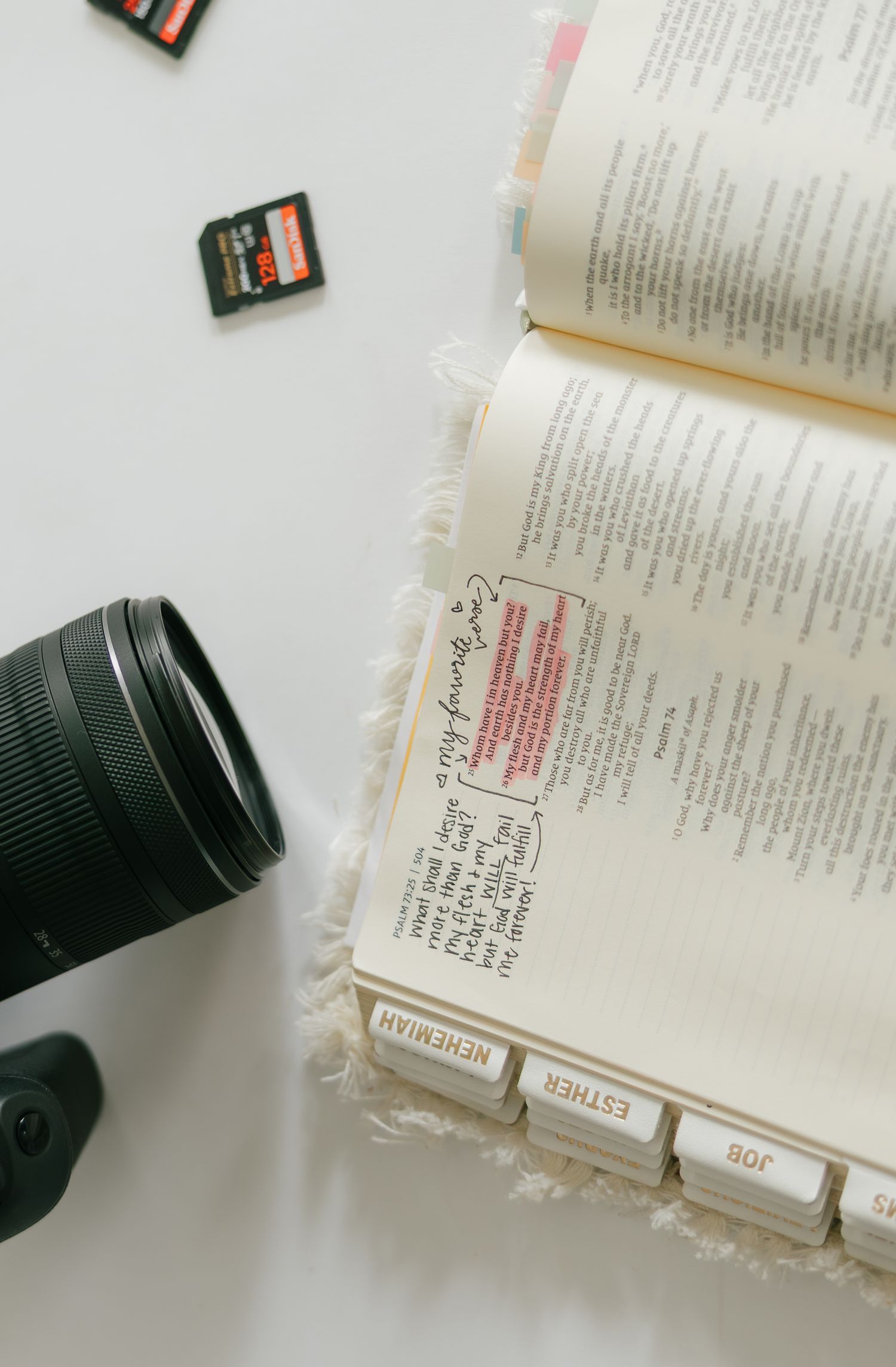 Open Bible with tabbed pages beside a camera lens and SD cards on a white surface.