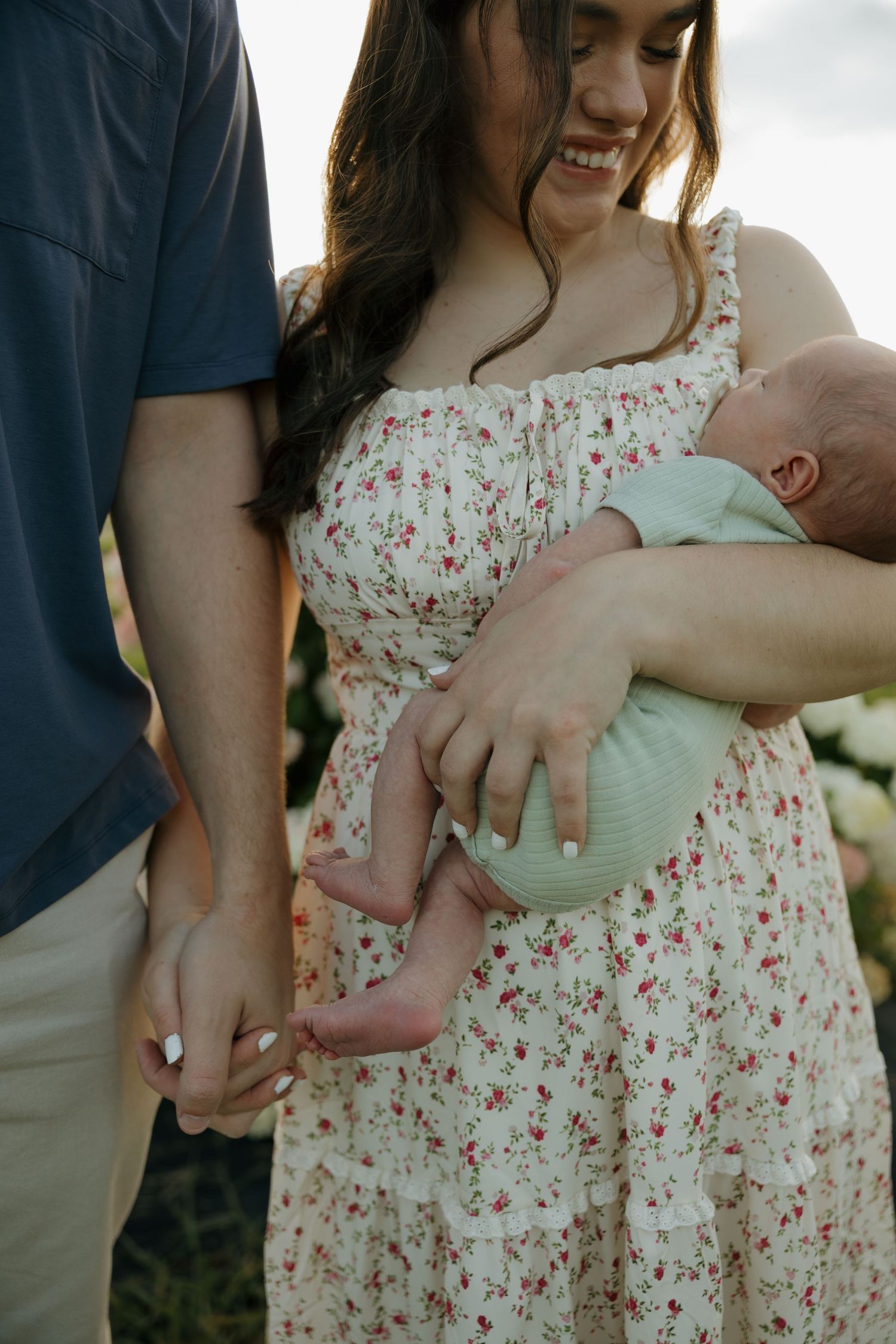 A smiling woman in a floral dress holds a newborn baby while a man holds her hand outdoors.