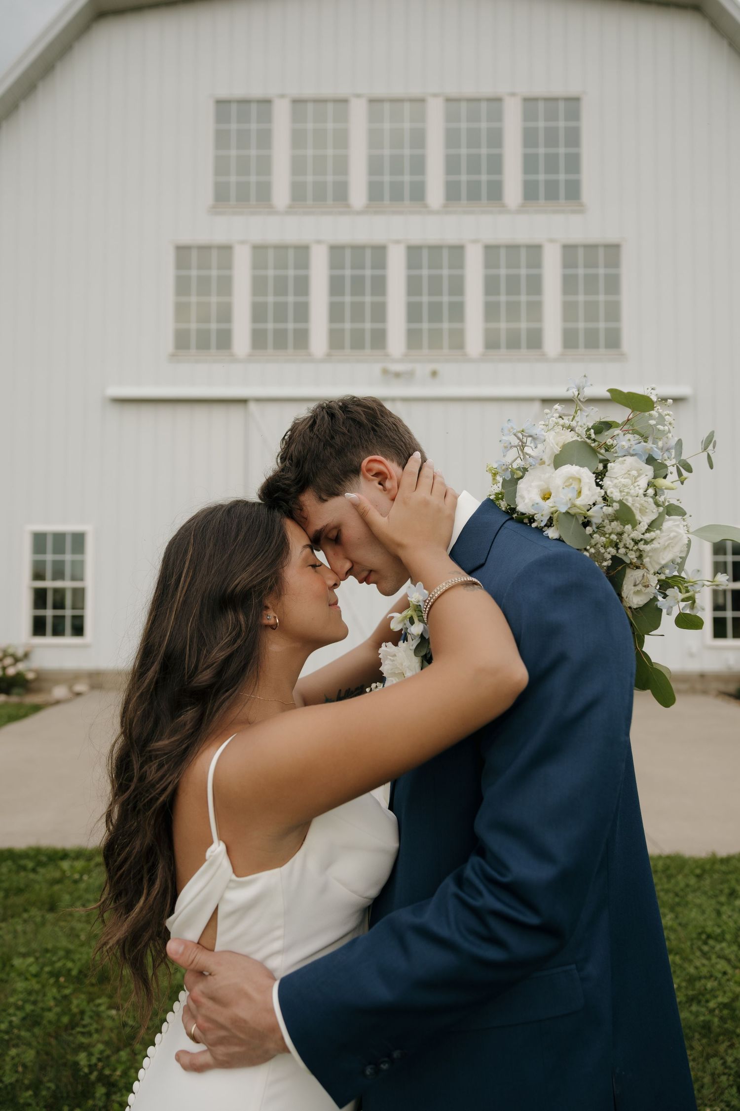 Bride and groom share a romantic forehead touch moment in front of a white barn wedding venue.