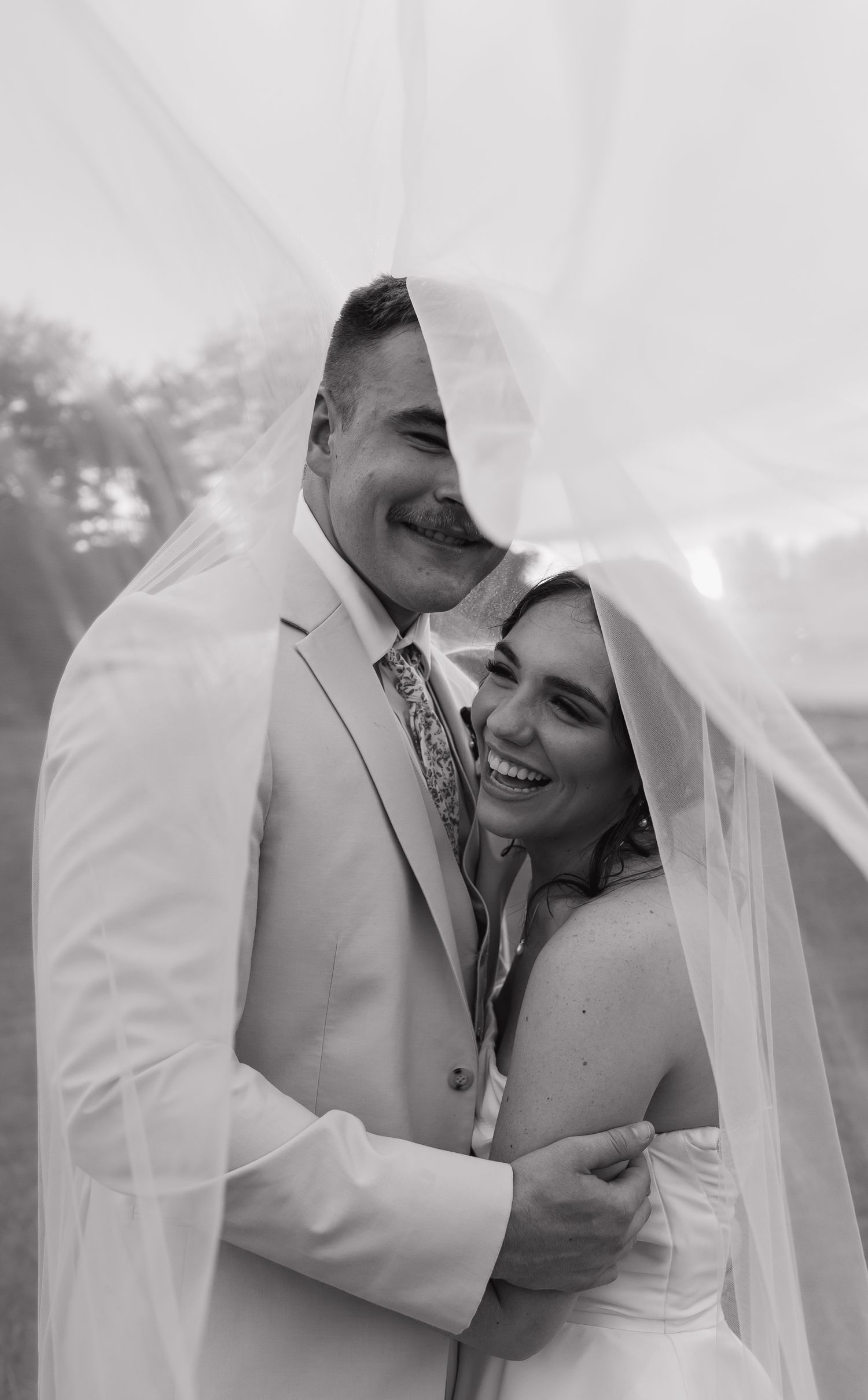 Black and white photo of a smiling bride and groom embracing under a flowing wedding veil.