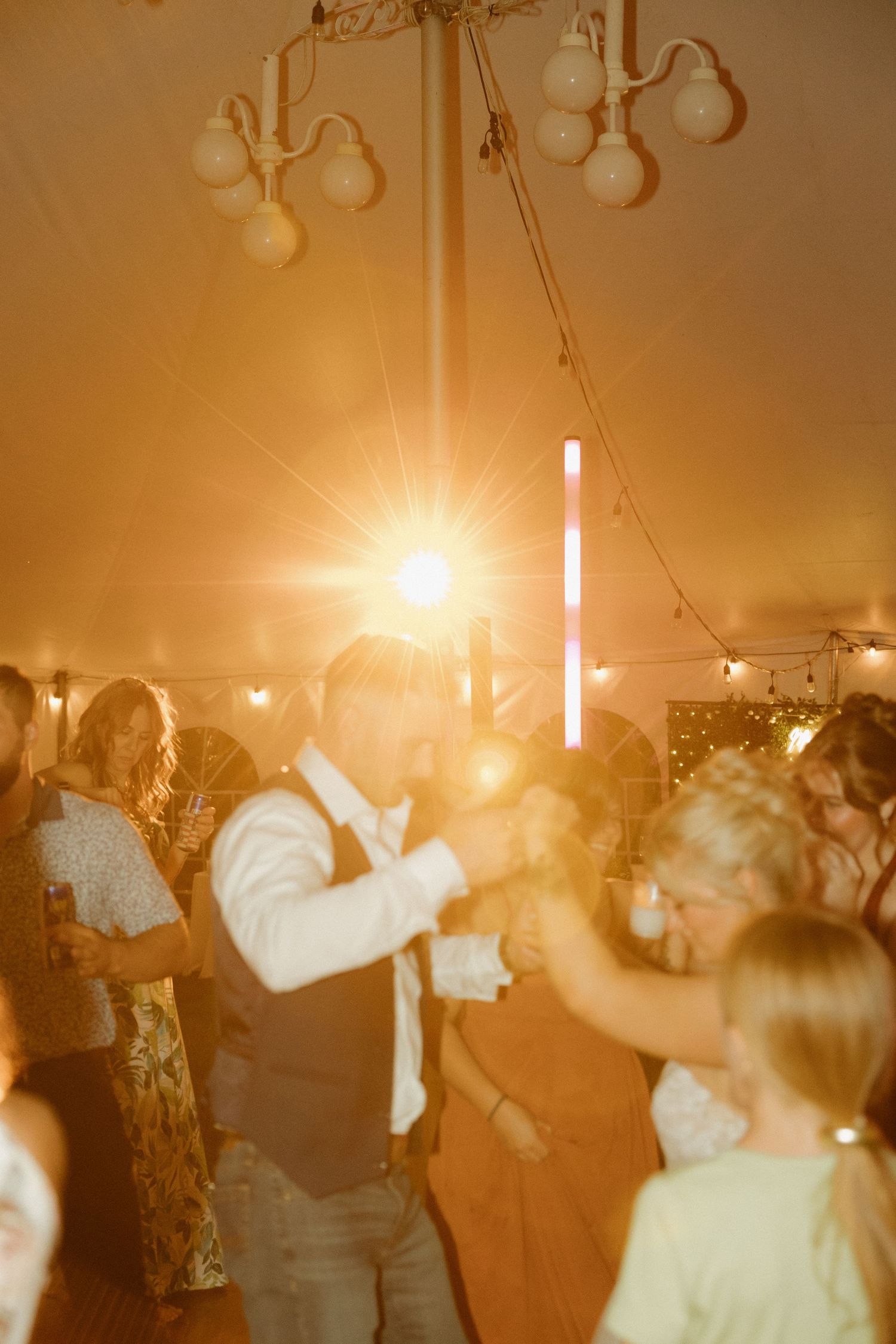 Wedding guests dance under warm glowing lights inside decorated tent at lively evening reception.