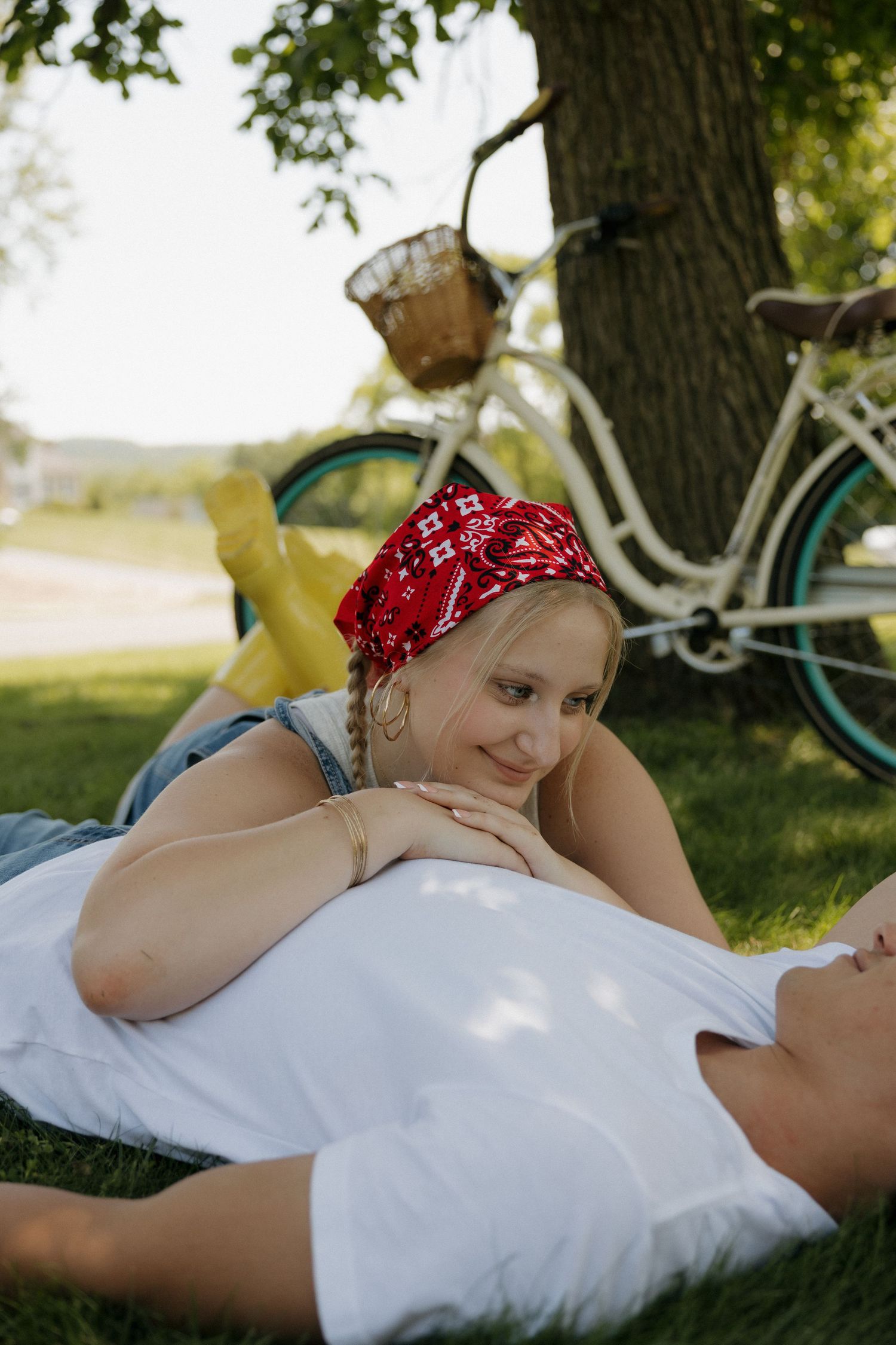 A couple relaxing on grass under a tree in a park, with vintage bicycles in the background on a sunny day.
