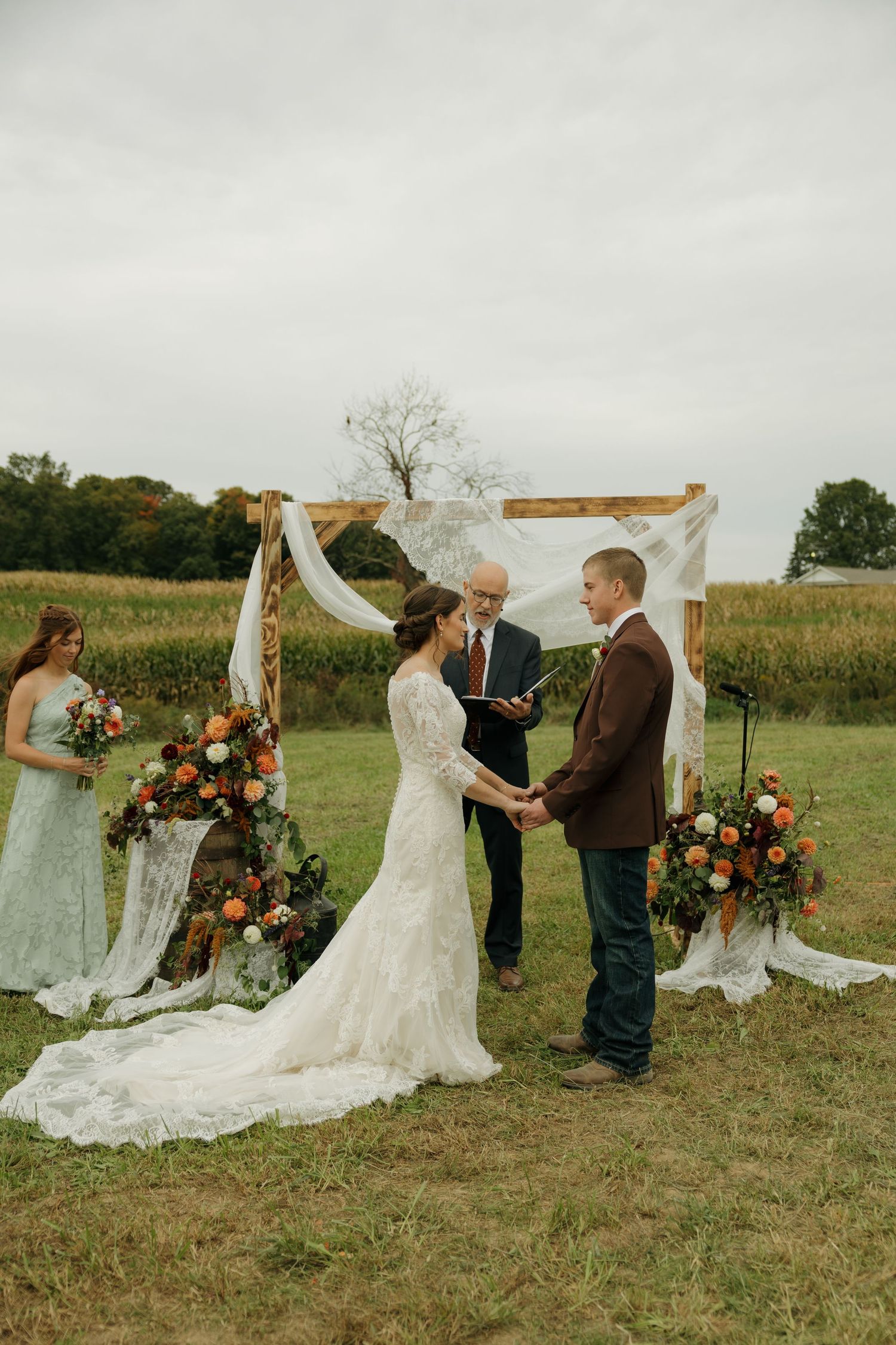 Bride and groom hold hands during an outdoor fall wedding ceremony under a draped wooden arch with orange floral arrangements.