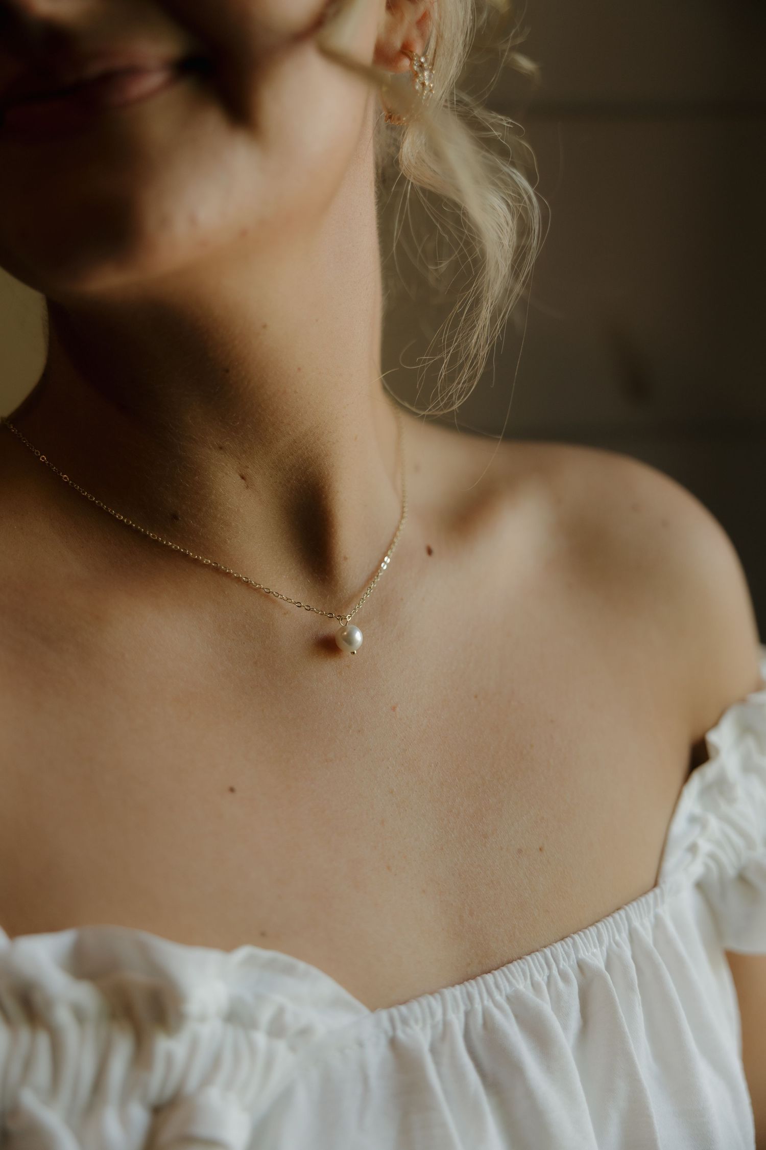 Woman wearing a delicate gold chain necklace with a single pearl pendant and white off-shoulder top.