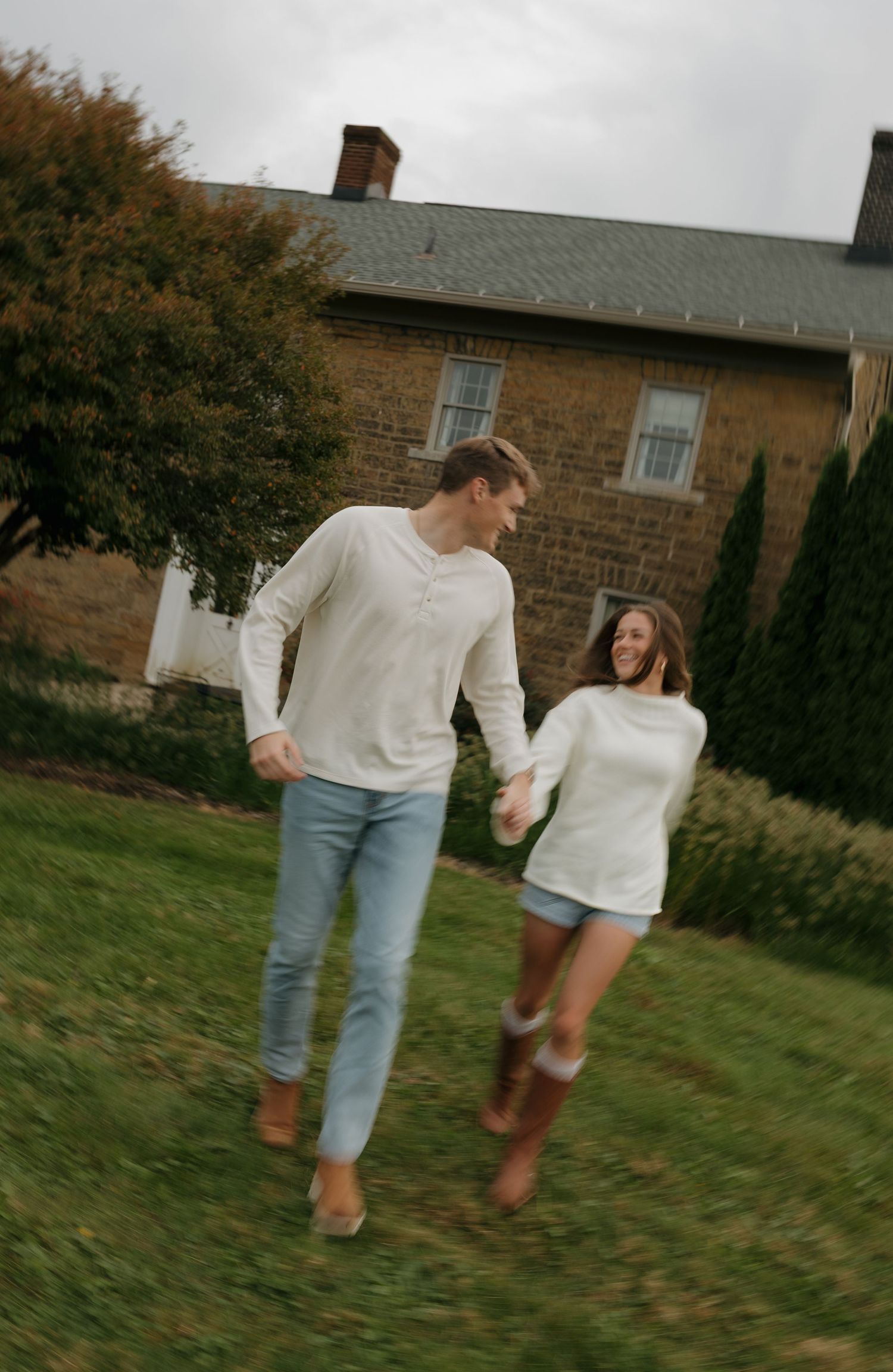A couple in matching white sweaters holds hands while running through a green lawn beside a rustic brick building.