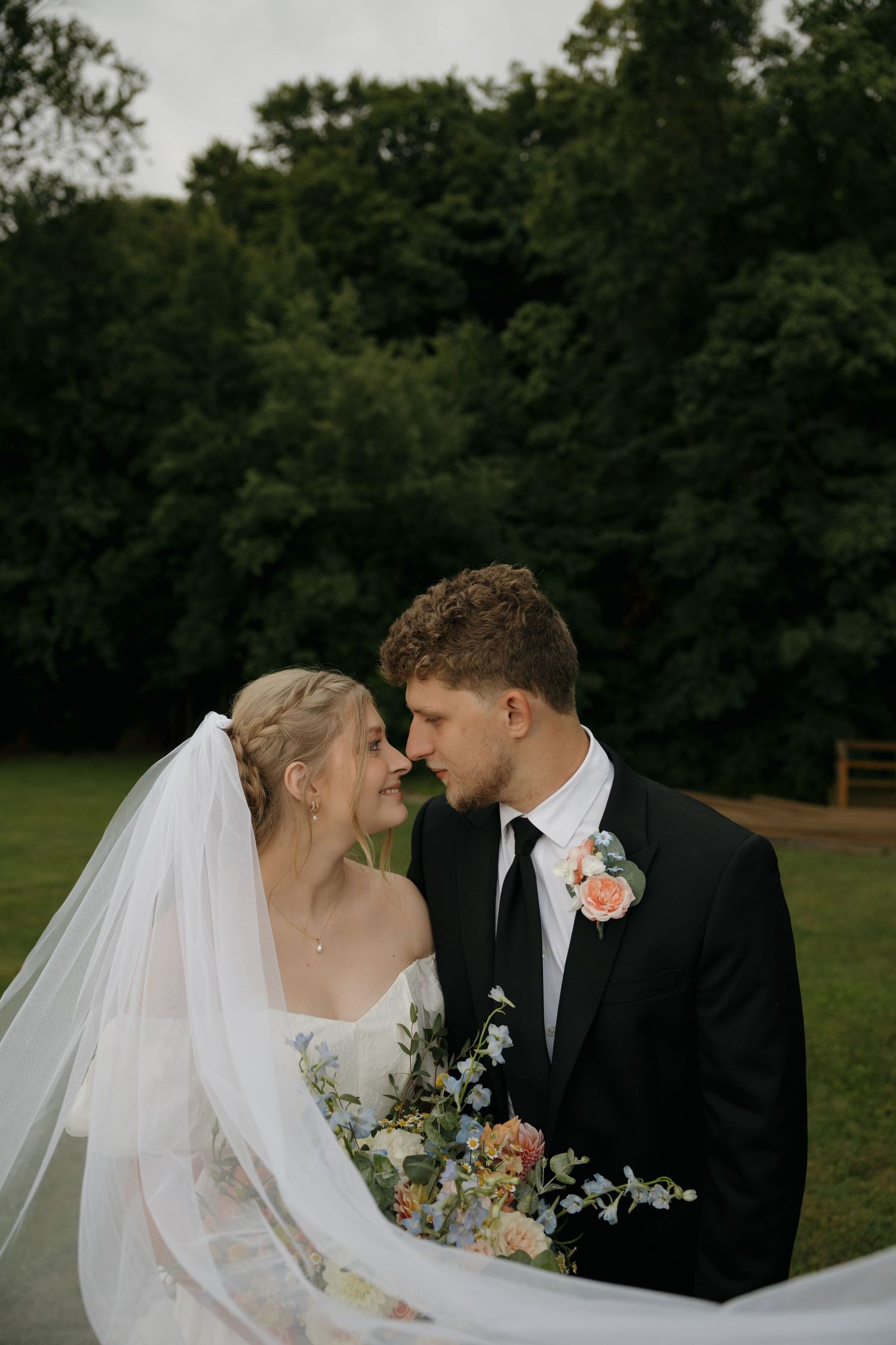 A bride and groom share a romantic moment outdoors, her veil flowing, as they lean in for a kiss at their wedding.