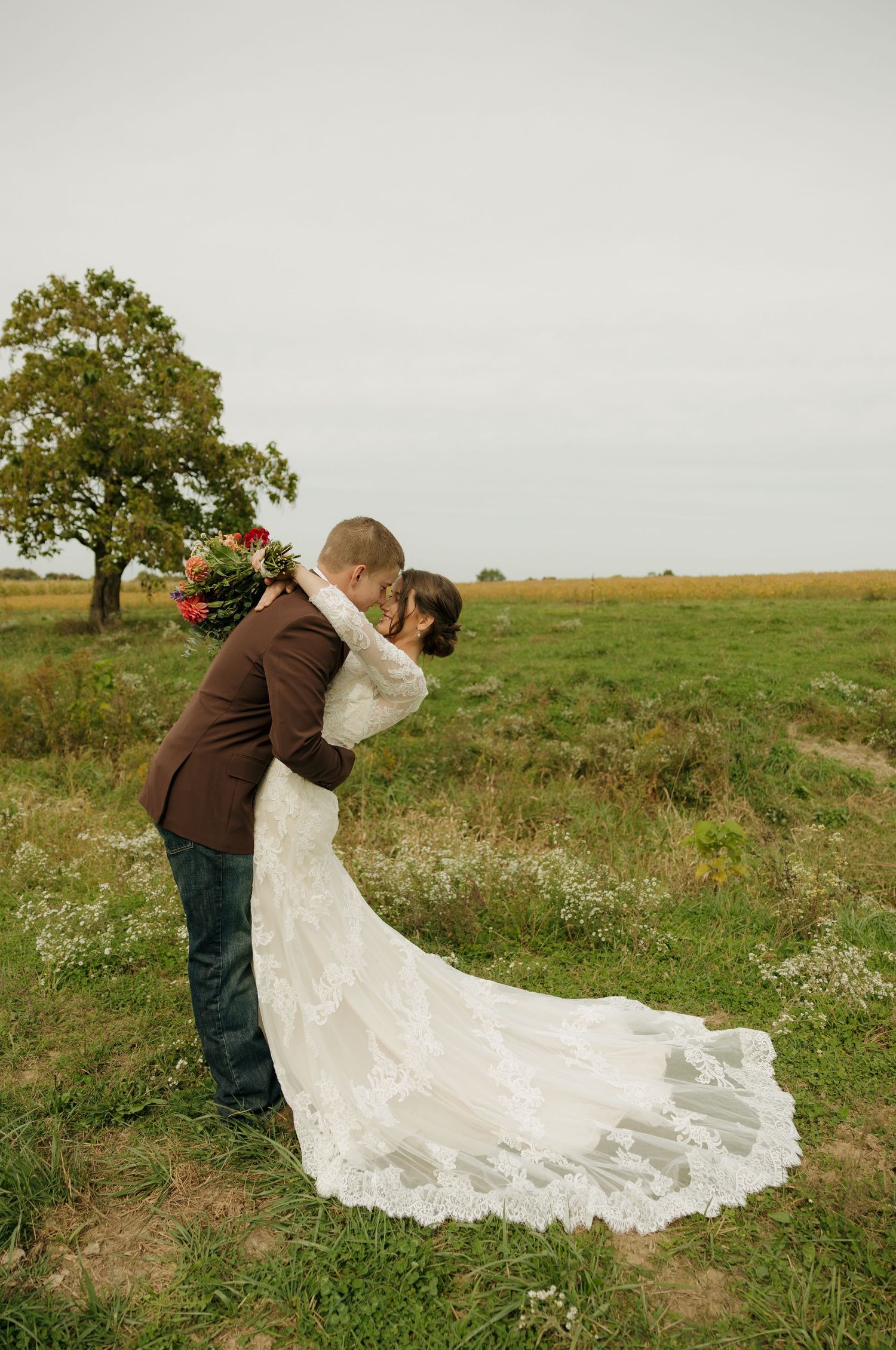 A groom in a brown jacket dips and kisses his bride in a lace wedding gown holding red flowers in a grassy field with a tree.