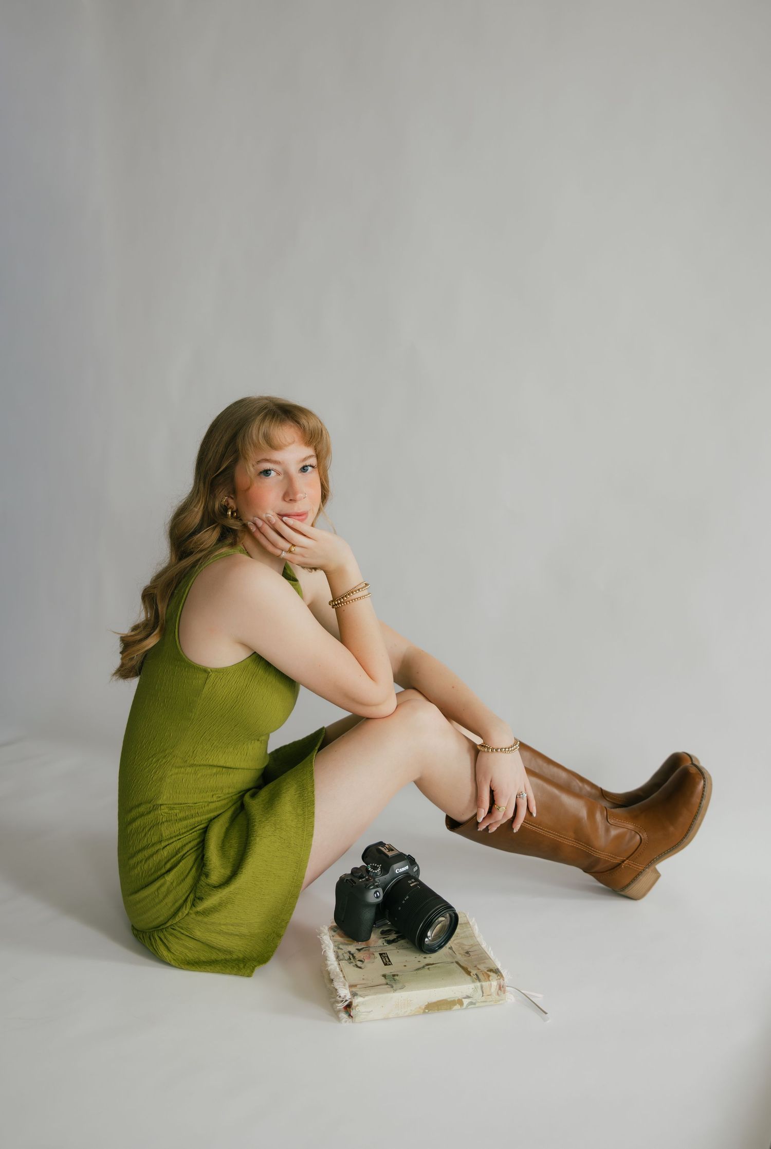 Young woman in green mini dress and brown boots sits on floor with camera and book.