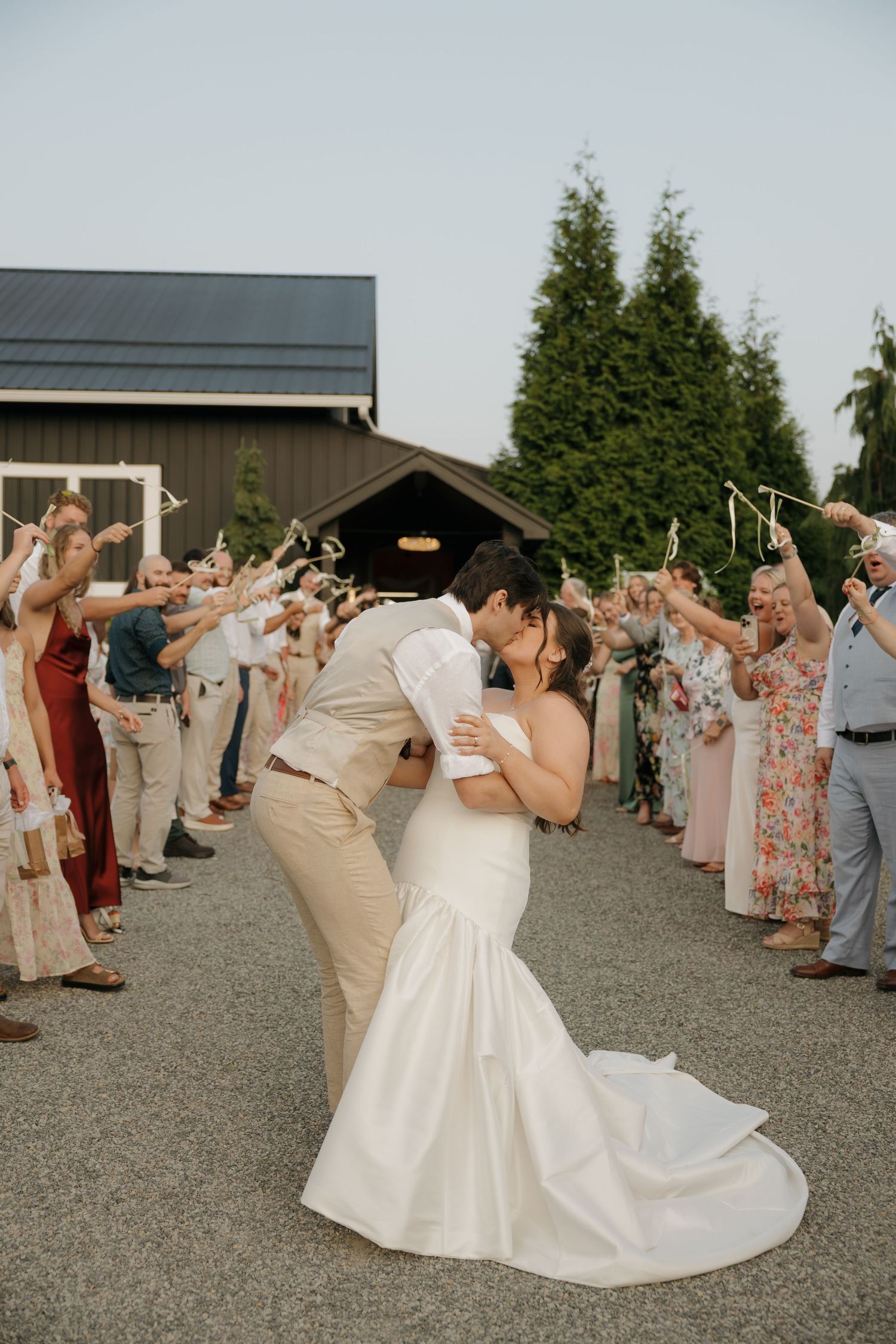 Bride and groom share a romantic kiss during their wedding sendoff, surrounded by cheering guests at an outdoor venue.