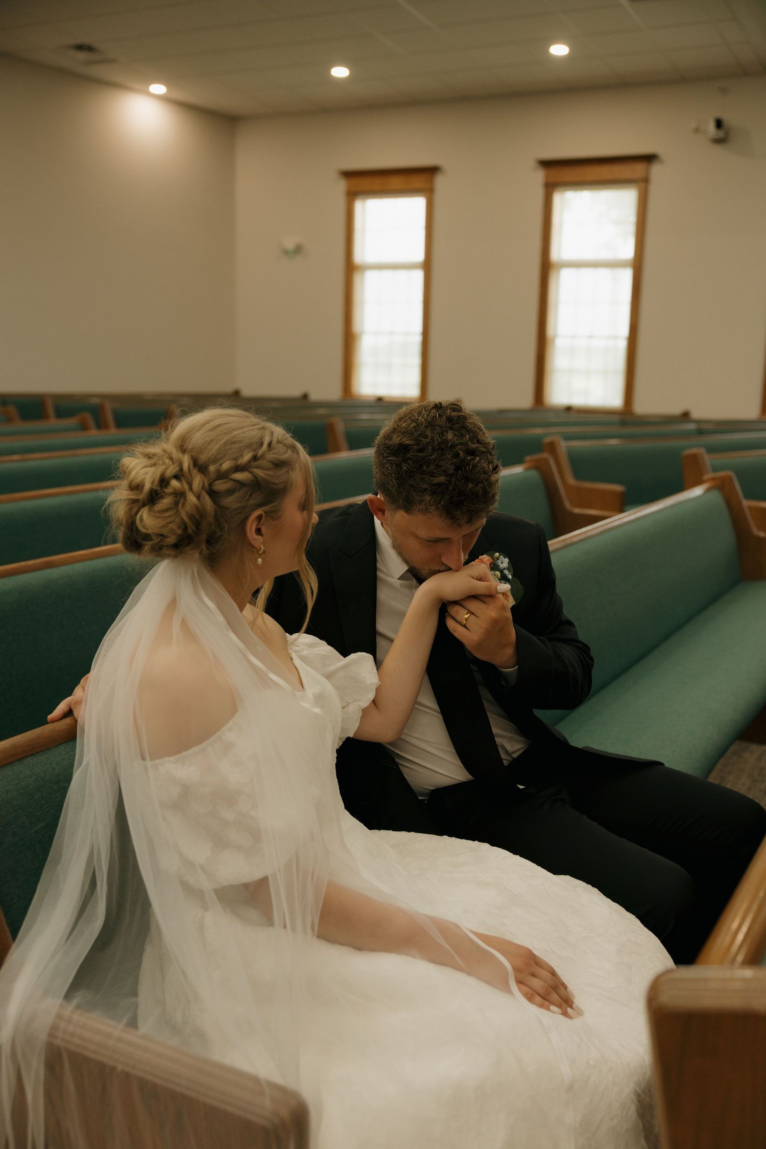 Bride and groom sit together in teal church pews, groom kissing bride's hand in a tender wedding moment.
