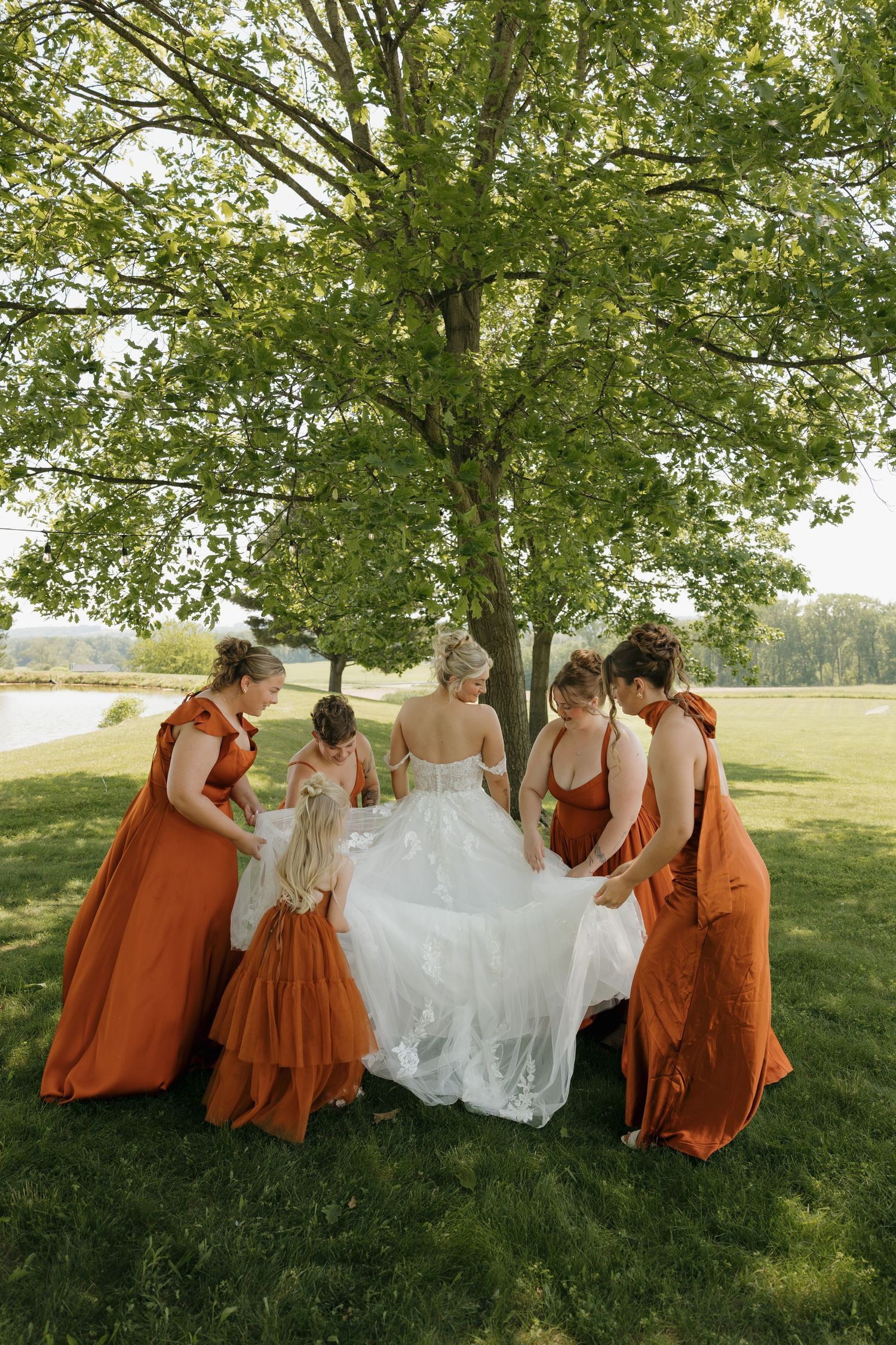 Bridesmaids in rust orange gowns gather around bride in white wedding dress under large green tree outdoors.