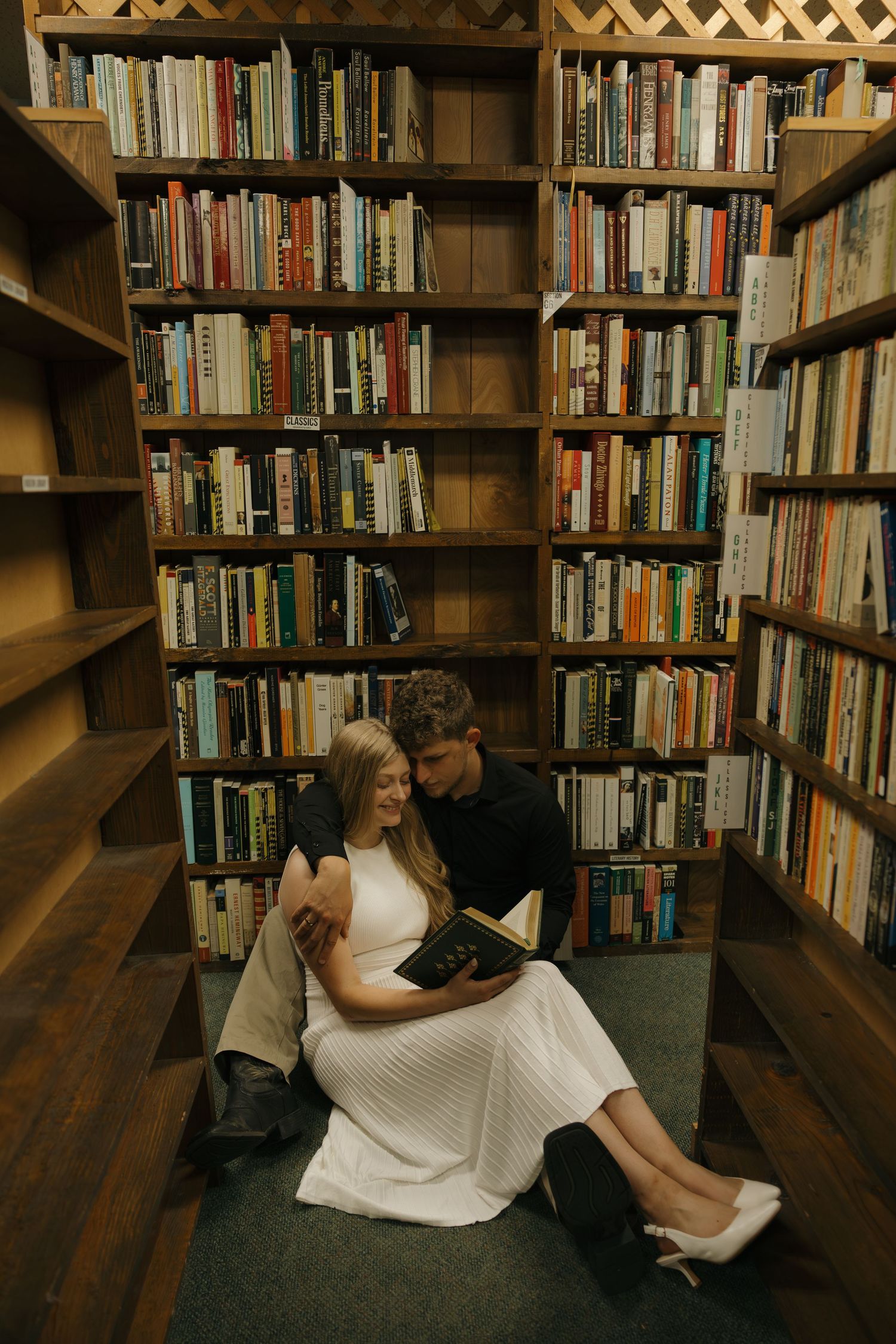 Couple sitting on floor reading a book together surrounded by tall wooden bookshelves in a cozy library.