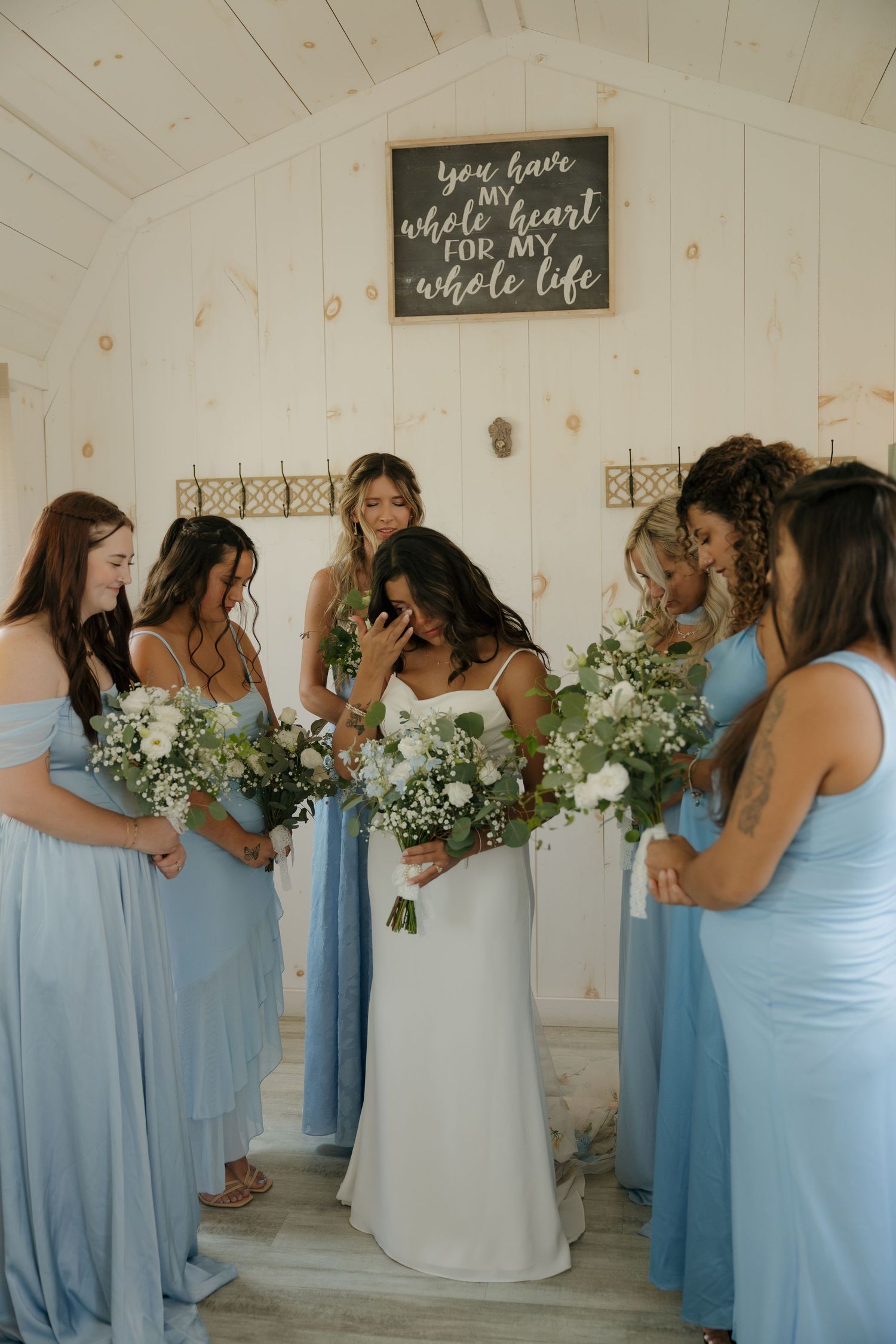Bride in white gown shares emotional moment with bridesmaids in blue dresses holding white bouquets indoors.