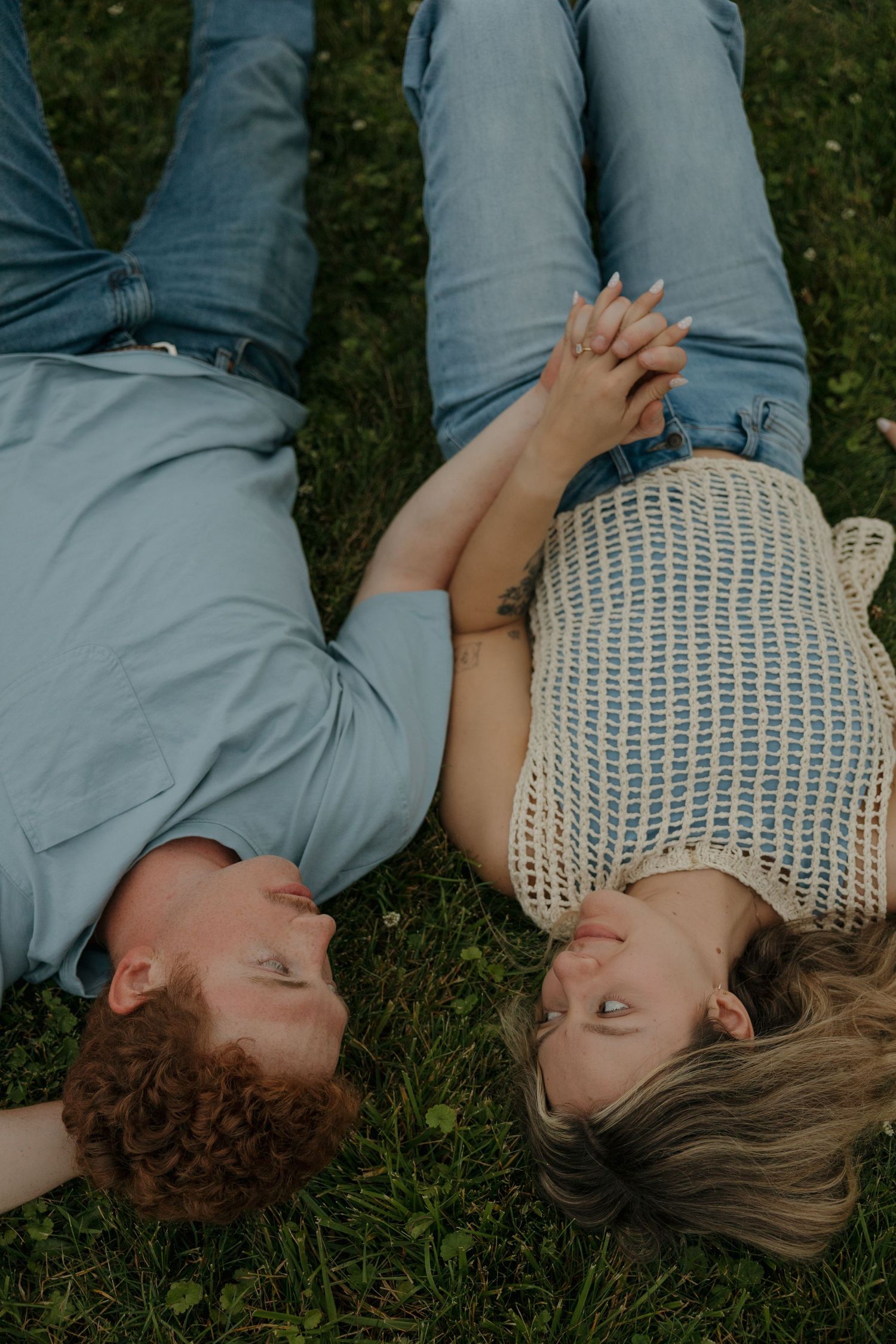 Couple lying on grass holding hands, smiling at each other in a romantic outdoor moment.