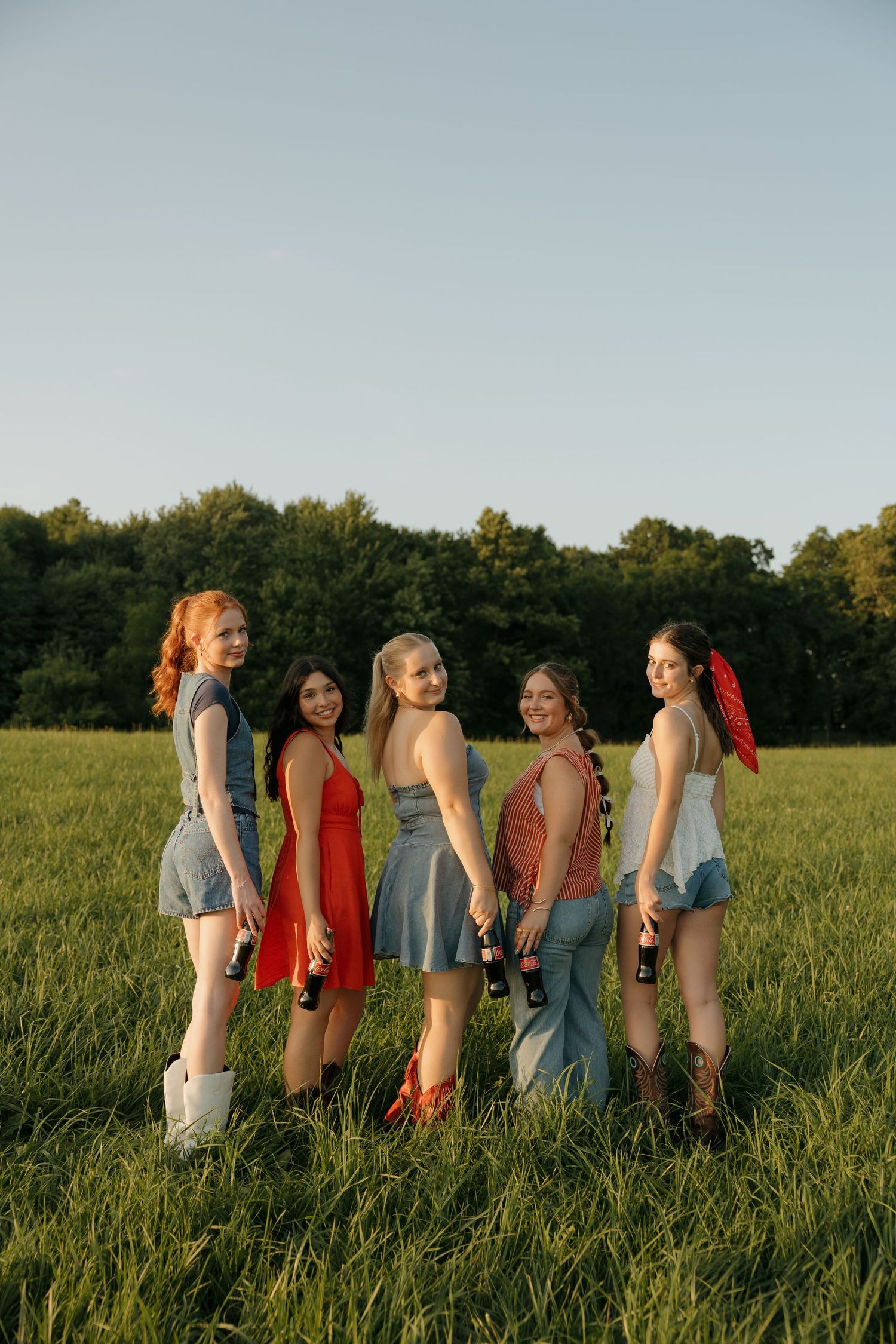Five young women in summer outfits holding bottles stand in a green field at golden hour with trees in the background.