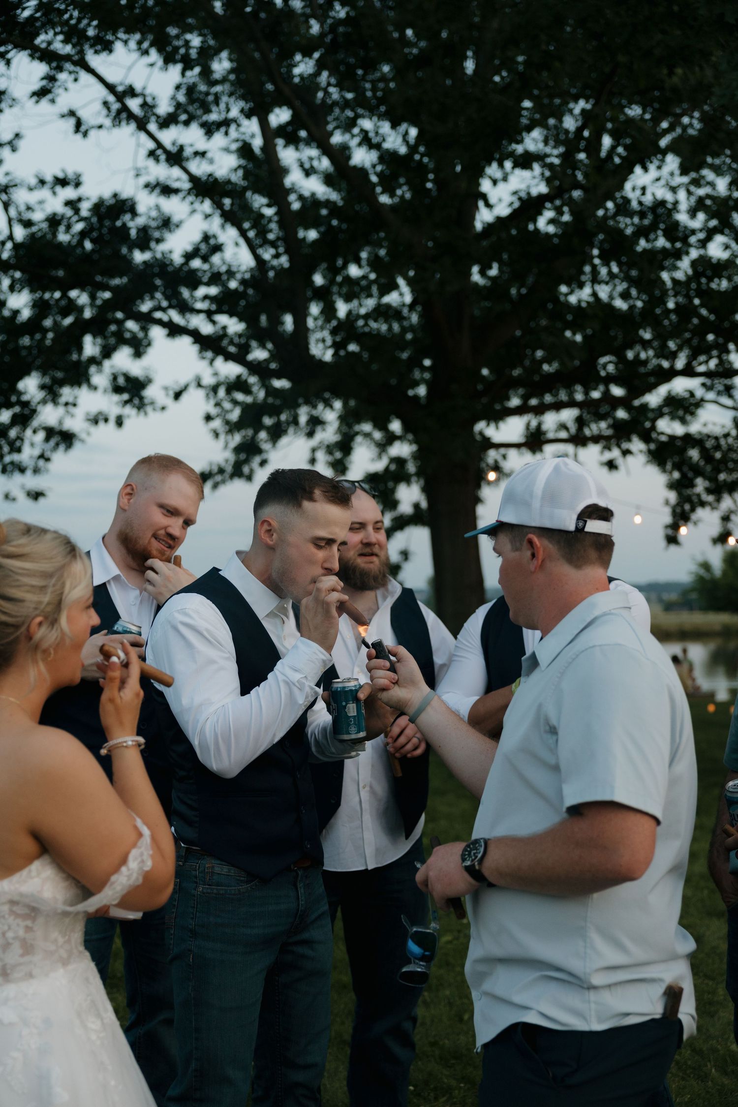 Groomsmen in white shirts and navy vests light cigars together outdoors at evening wedding reception.