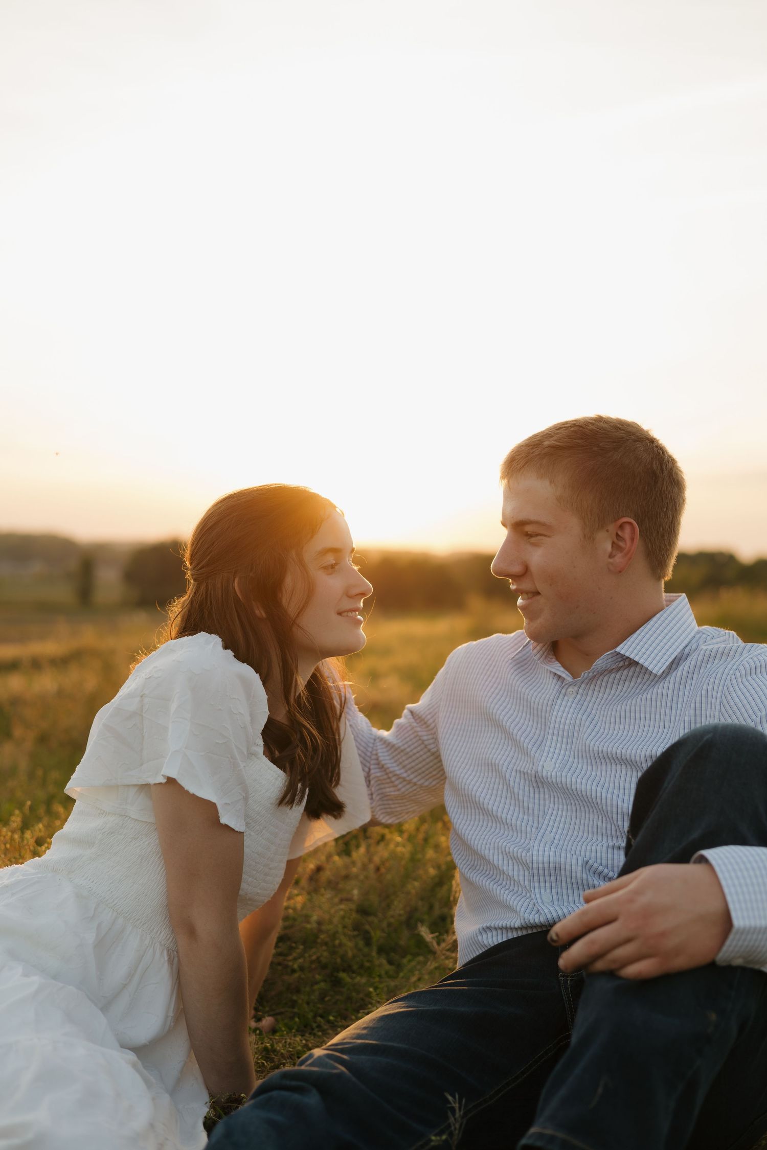 Couple sitting in golden sunset field, smiling at each other, woman in white dress, man in striped shirt, romantic outdoor photo.