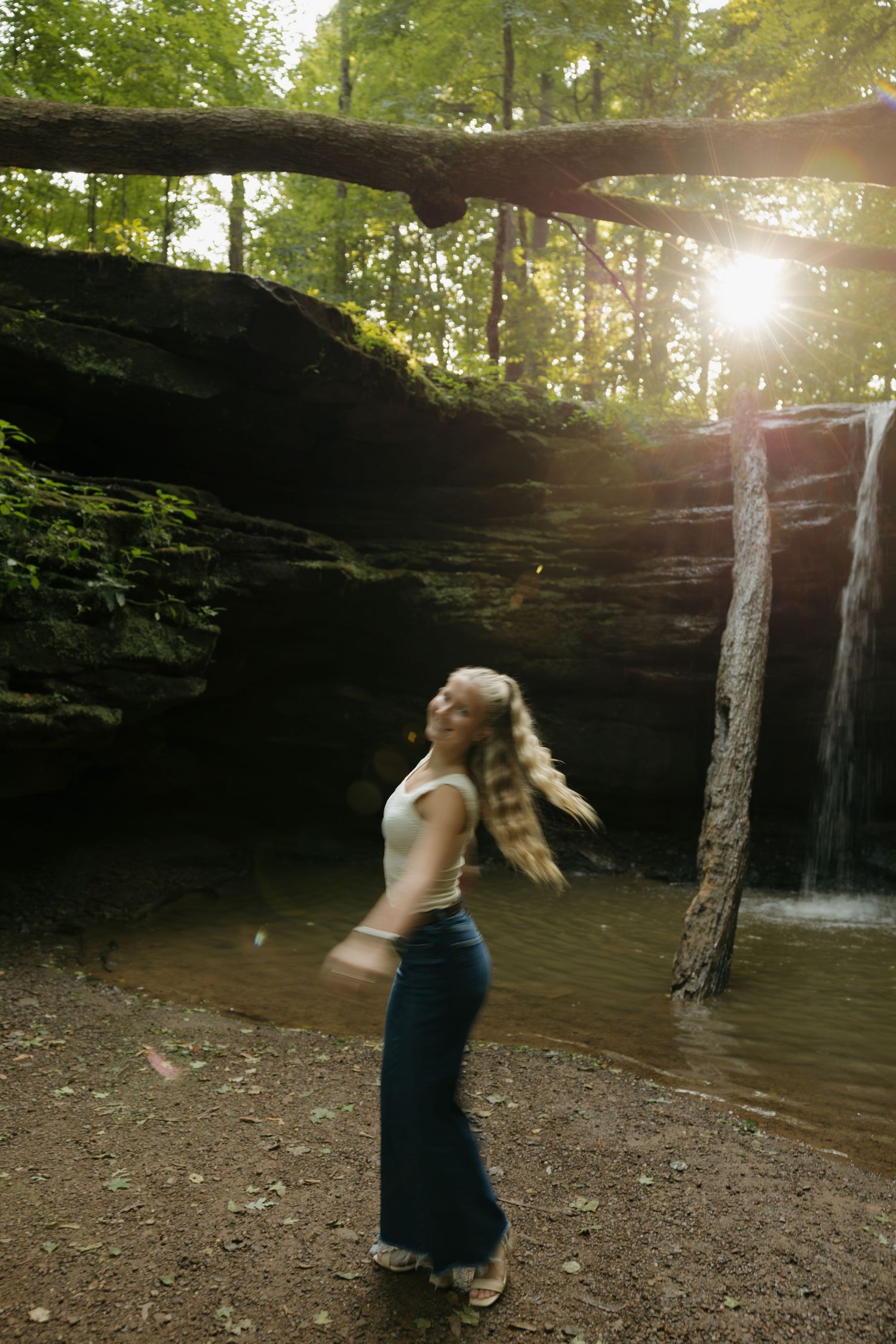 Woman with flowing blonde hair twirling near a sunlit waterfall in a lush forest wearing jeans and white top.