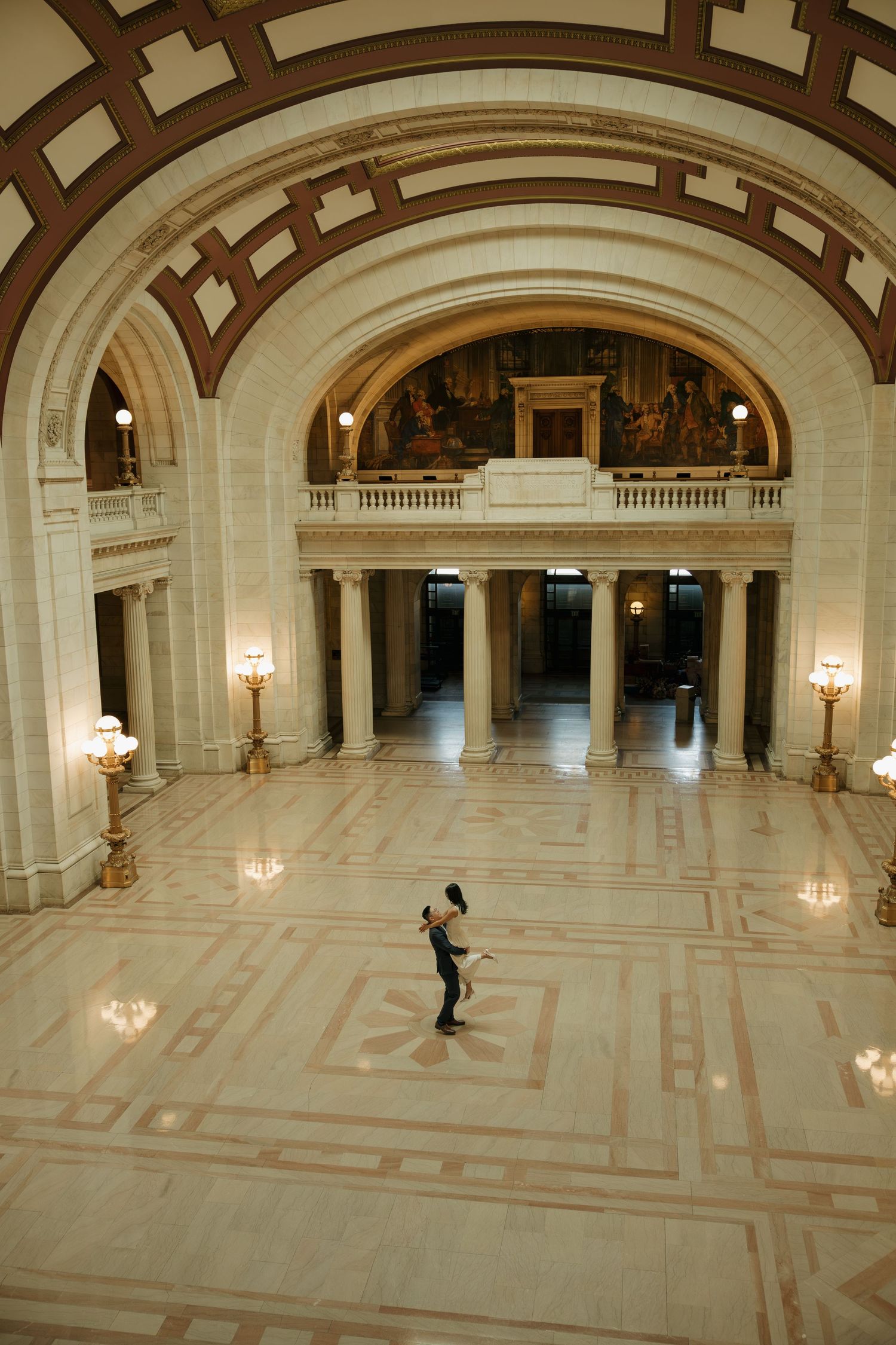 Two people dance in the grand marble rotunda of a neoclassical building with arched ceilings and ornate columns.
