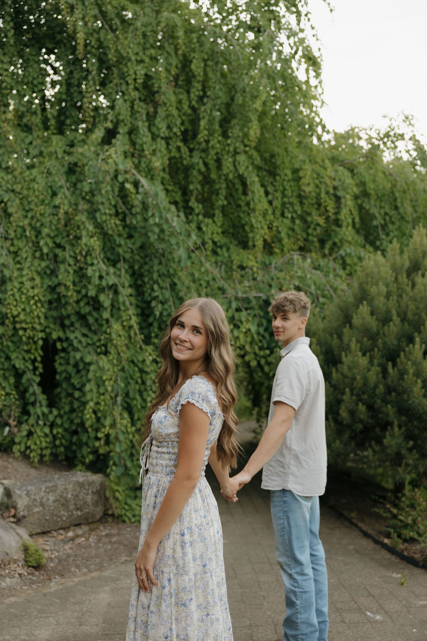 A smiling couple holds hands on a garden path, woman in floral dress, man in white shirt and jeans, lush green trees behind.