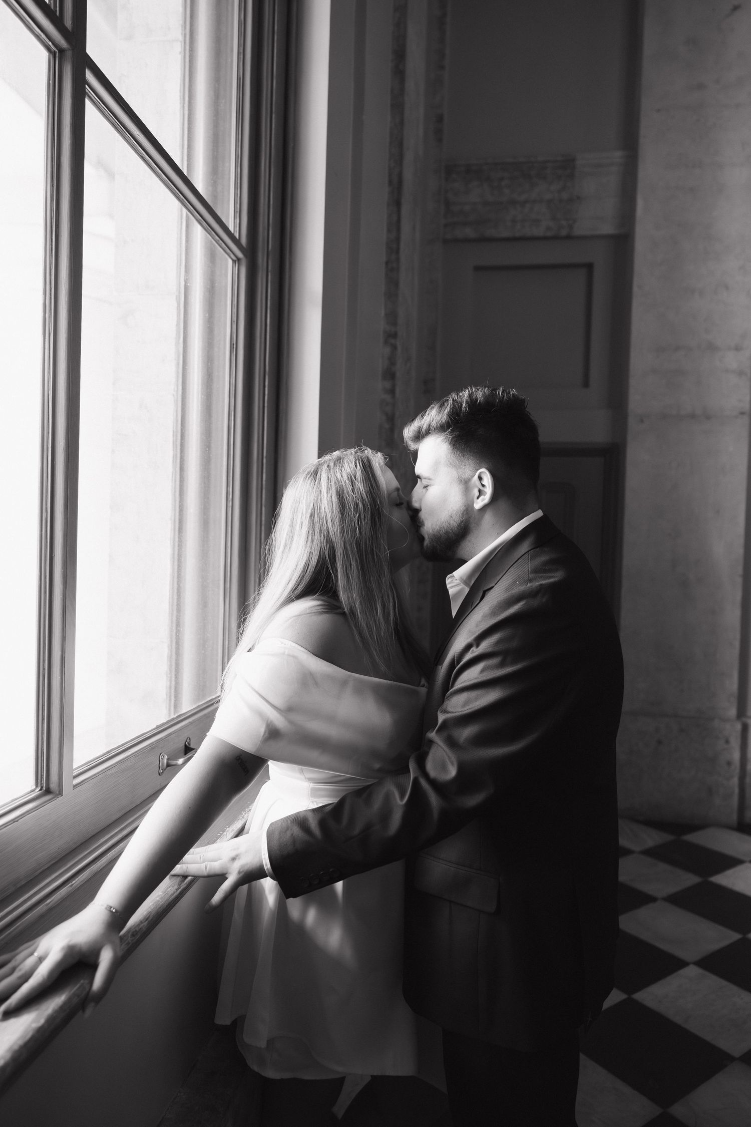 Black and white photo of a couple sharing a romantic kiss by a large window with checkered floor.