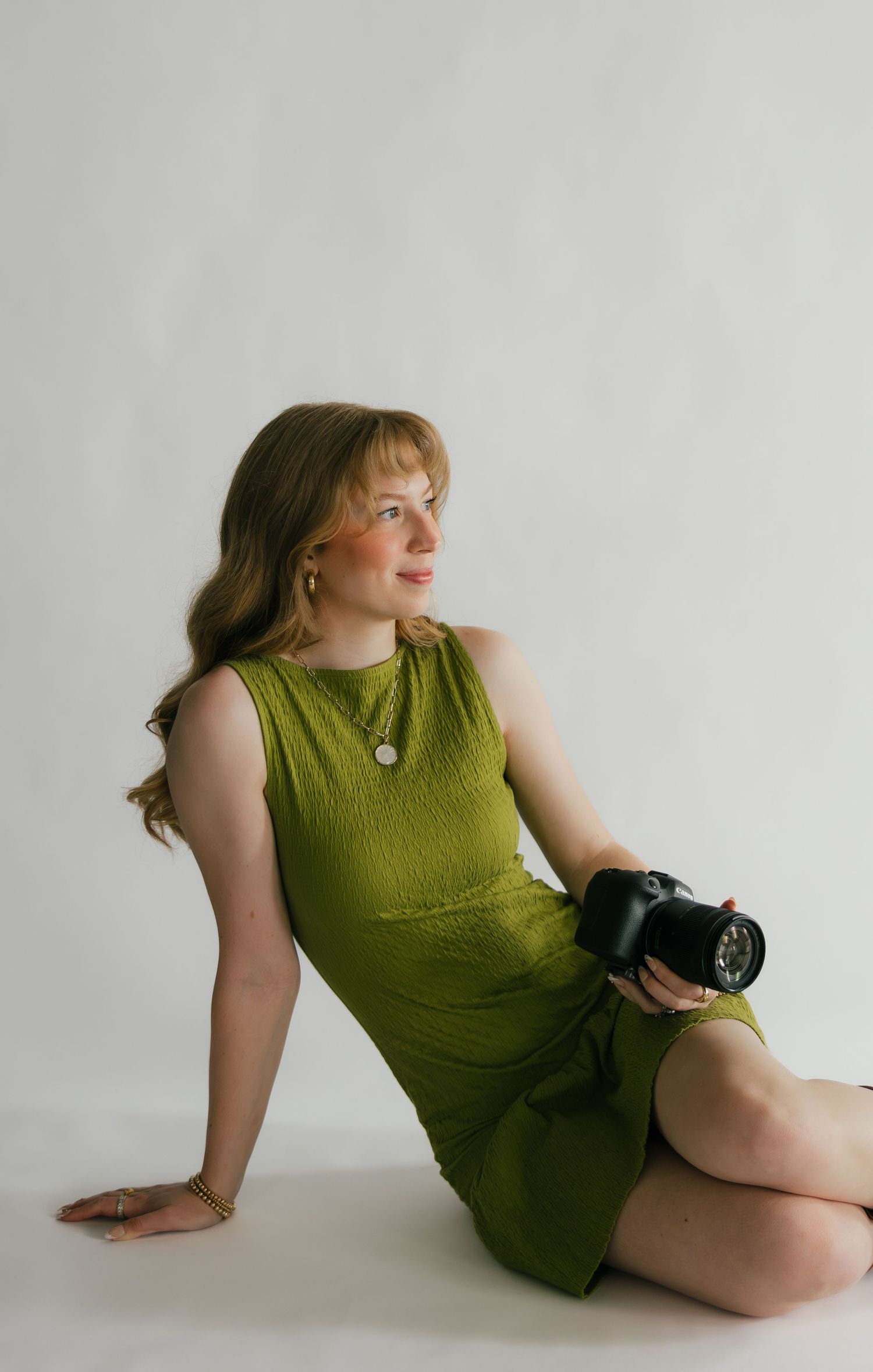 A young woman in a green mini dress sits on a white studio floor holding a DSLR camera, looking to the side.