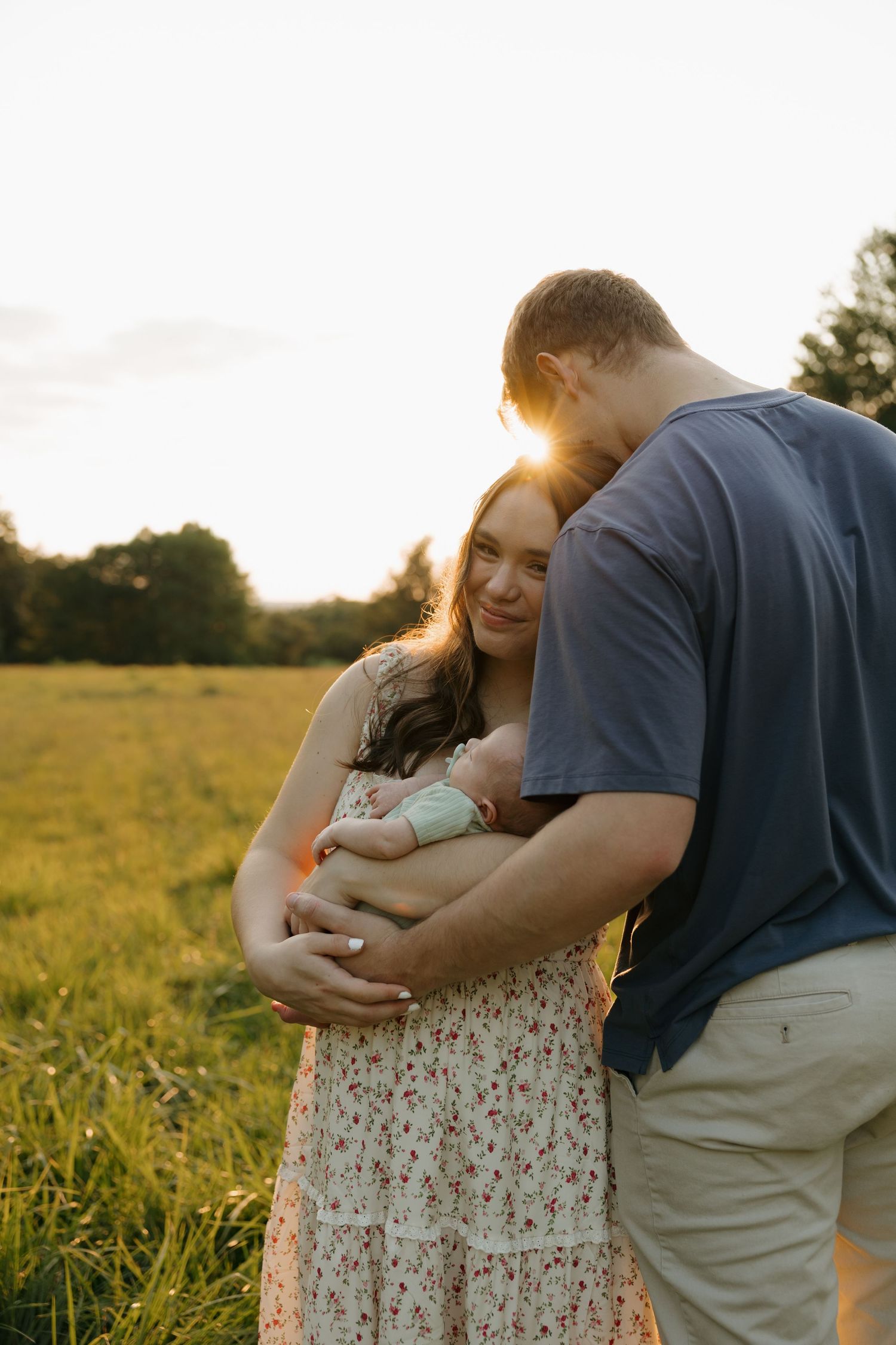 A couple embraces their newborn baby in a golden sunset field, capturing a tender family moment outdoors.