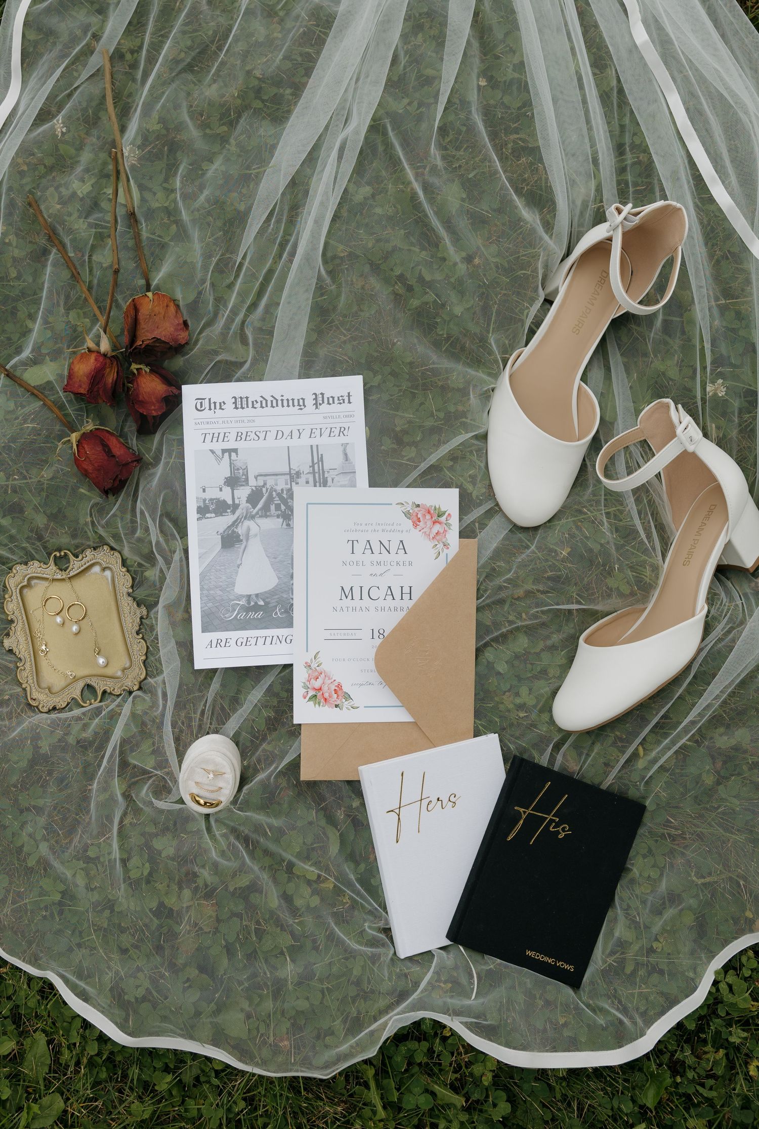 Elegant wedding flat lay with white heels, floral invitations, veil, dried roses, and His & Hers vow books on grass.