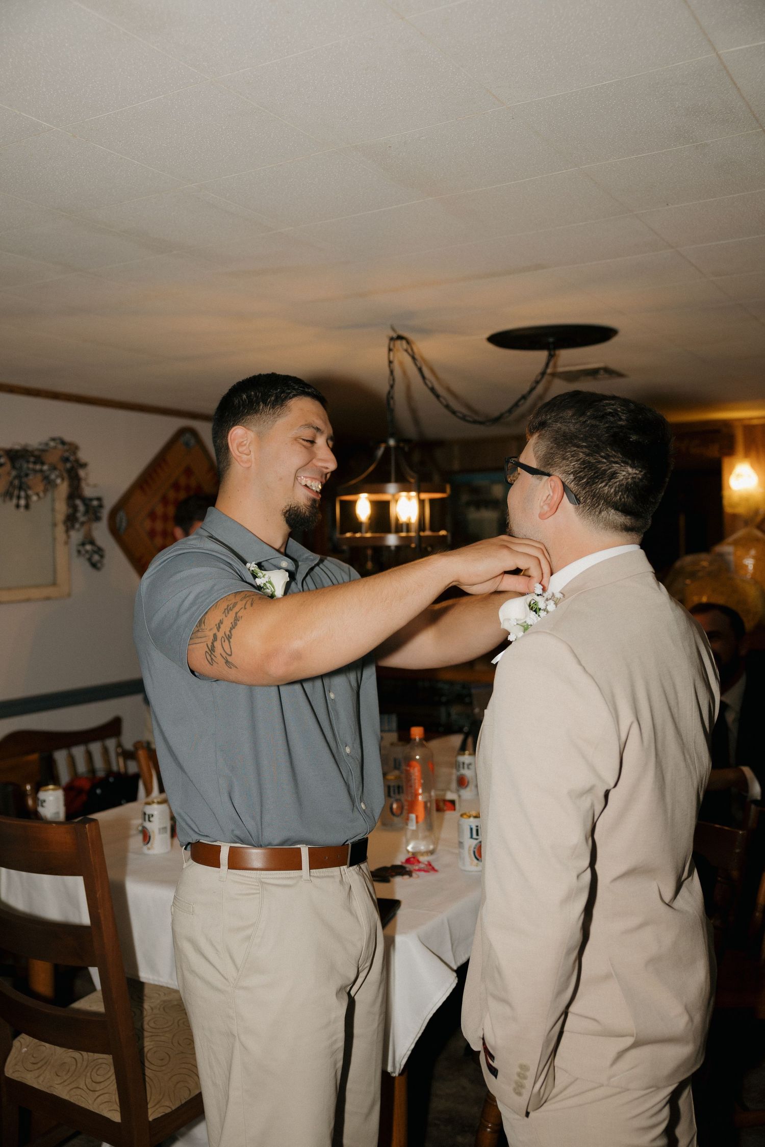 Smiling groomsman pinning a boutonniere on the groom in a beige suit at a wedding reception.