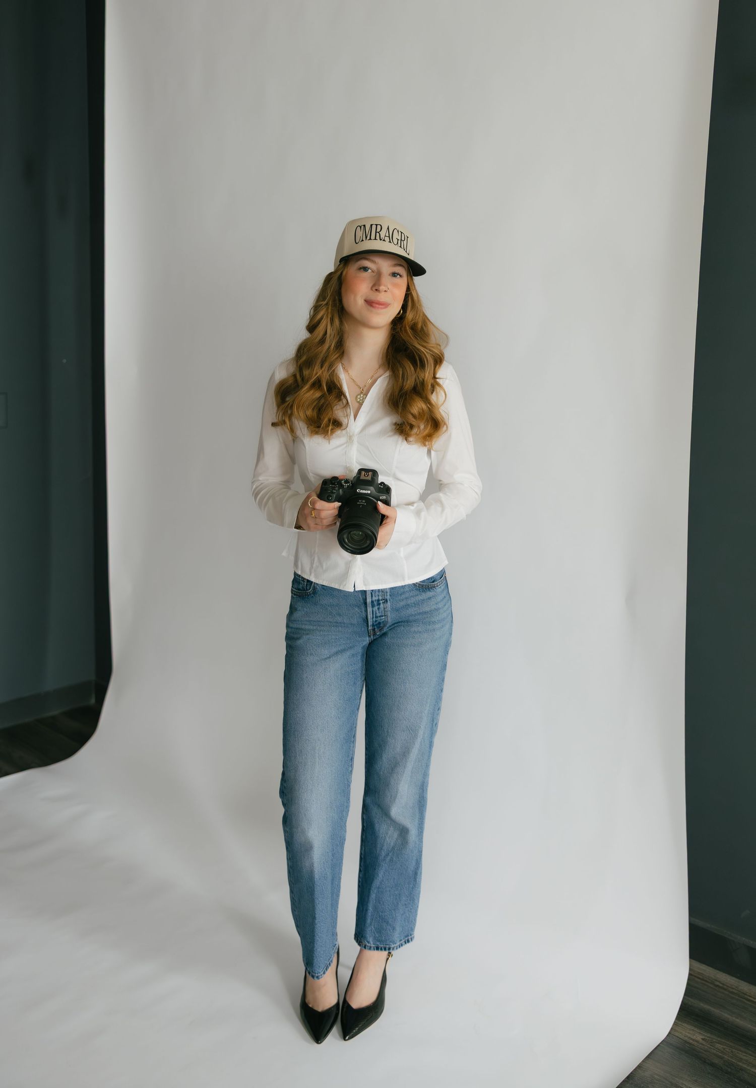 Young woman holding a camera in a white button-up shirt, jeans, heels, and a cap against a white backdrop.