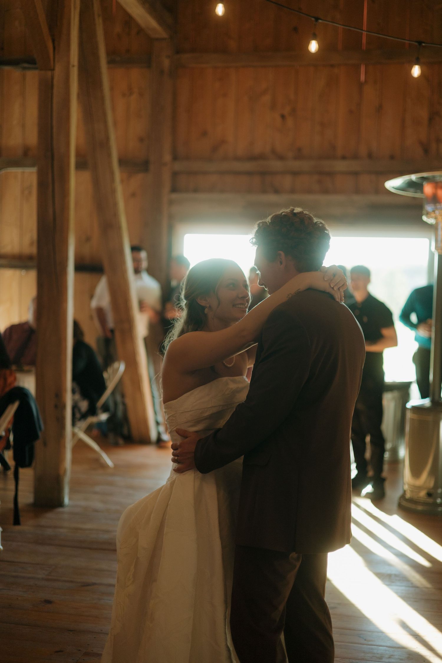 Bride and groom share a first dance in a rustic wooden barn as warm sunlight streams in and guests look on.