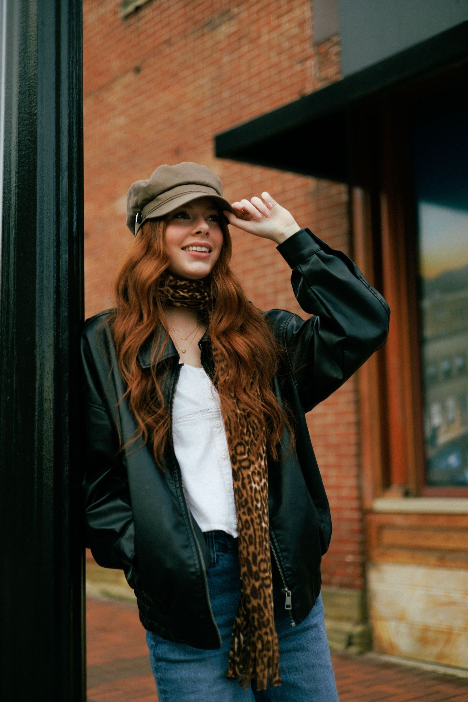 A casual street style photo featuring a newsboy cap, leopard scarf, and bomber jacket against brick architecture.