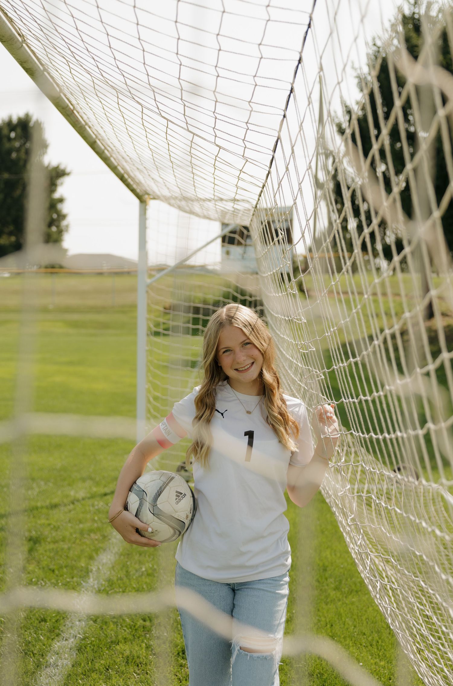 A smiling teenage girl in a soccer jersey holds a ball while leaning against a goal net on a green field.