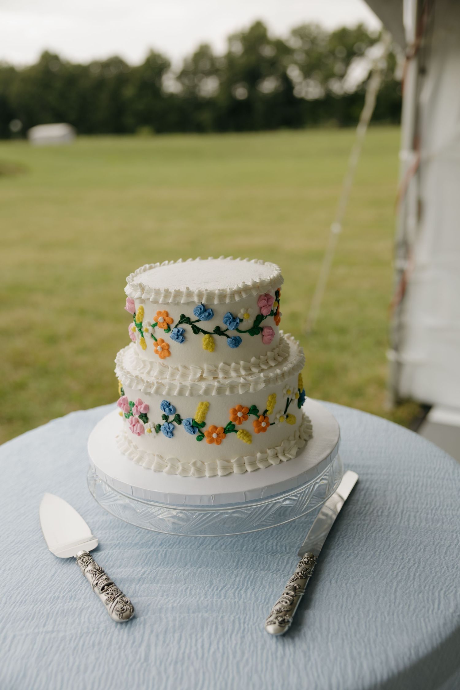 Two-tier white wedding cake with colorful floral piping, displayed on a blue tablecloth with silver cake server and knife.