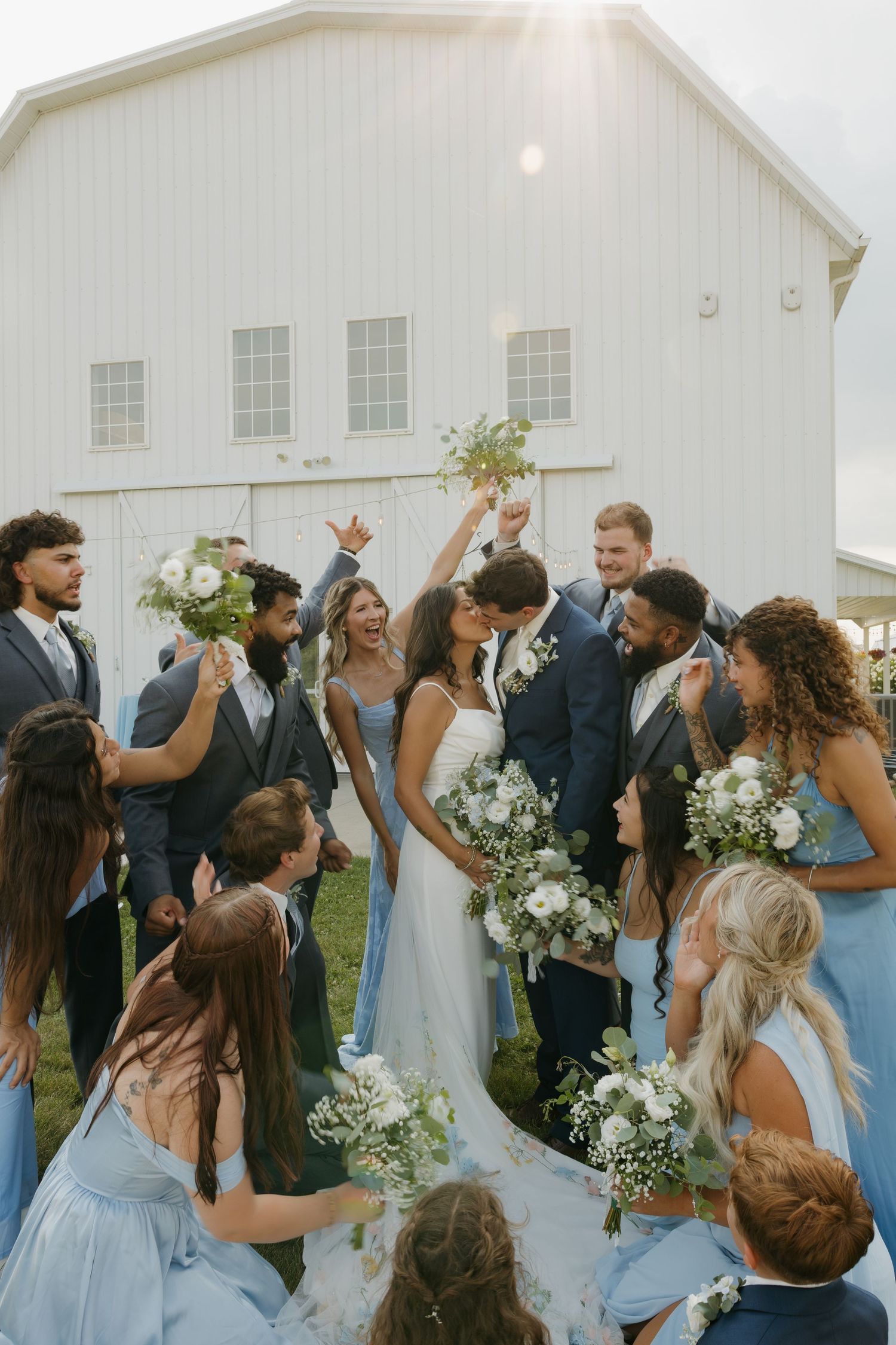 Wedding party celebrates with the newlyweds kissing in front of a sunlit white barn venue.