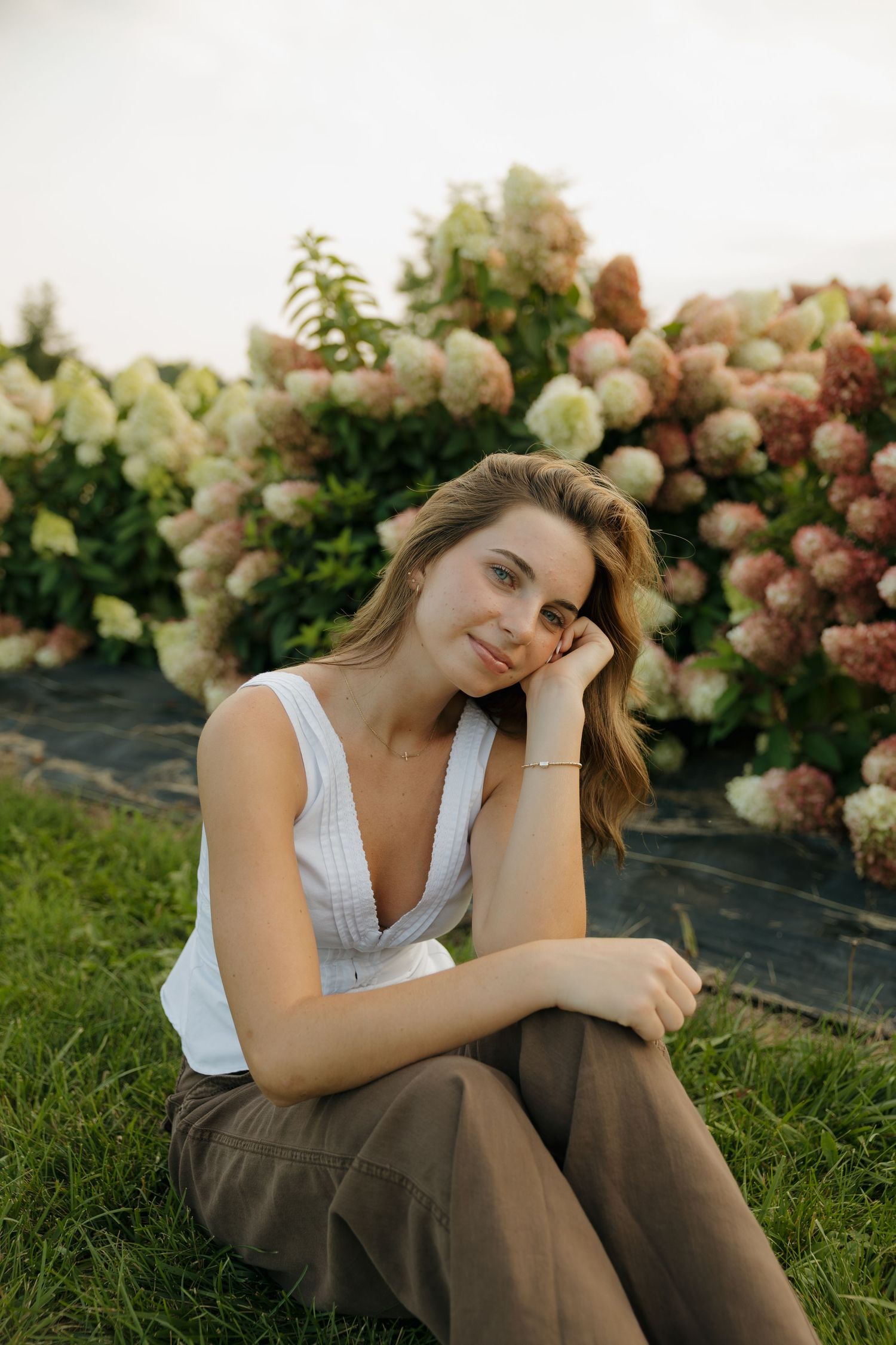 Young woman in white tank top and brown pants sitting on grass in front of blooming hydrangea bushes.