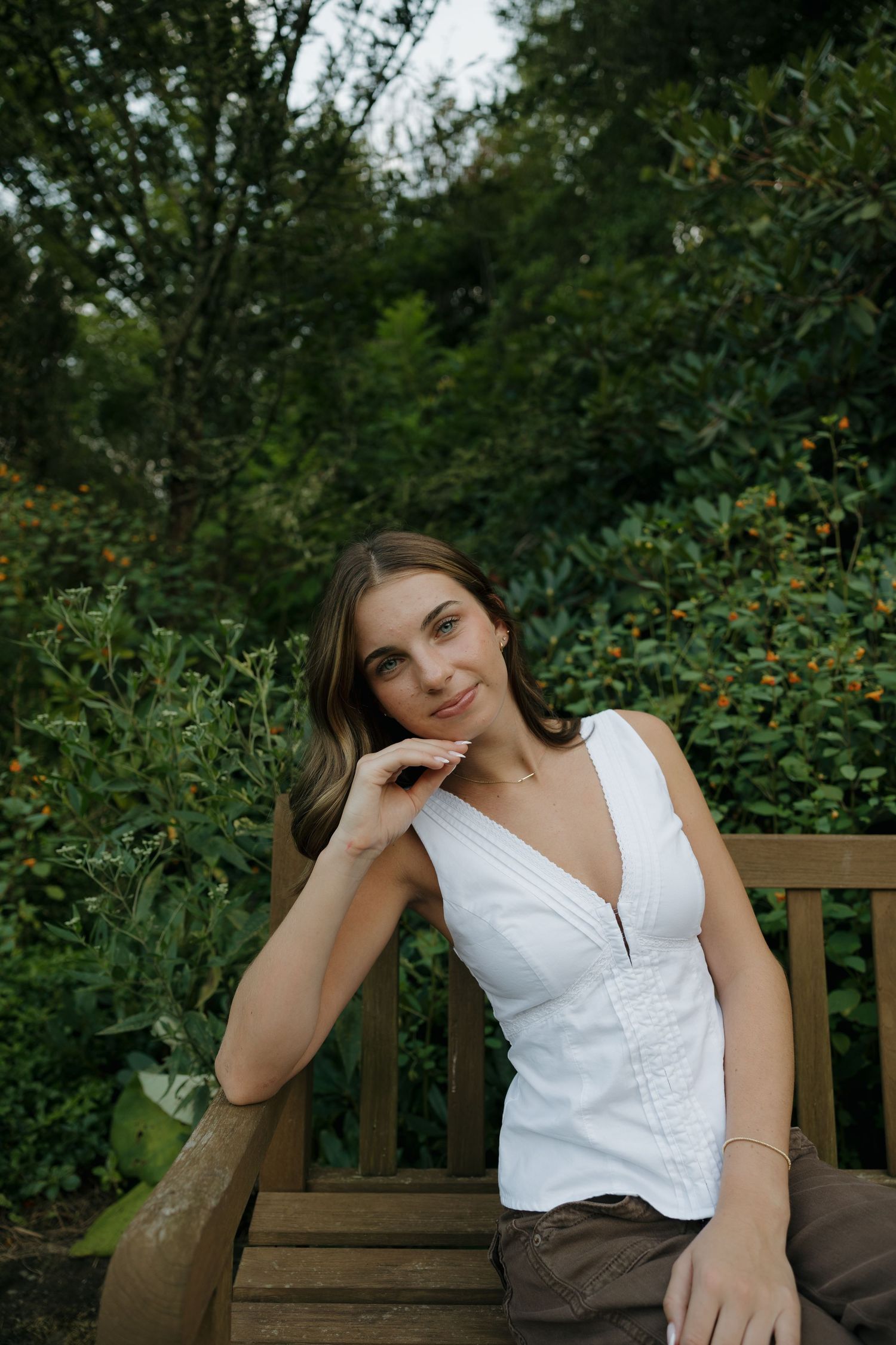 Young woman in white sleeveless top sitting on wooden bench surrounded by lush green garden foliage.