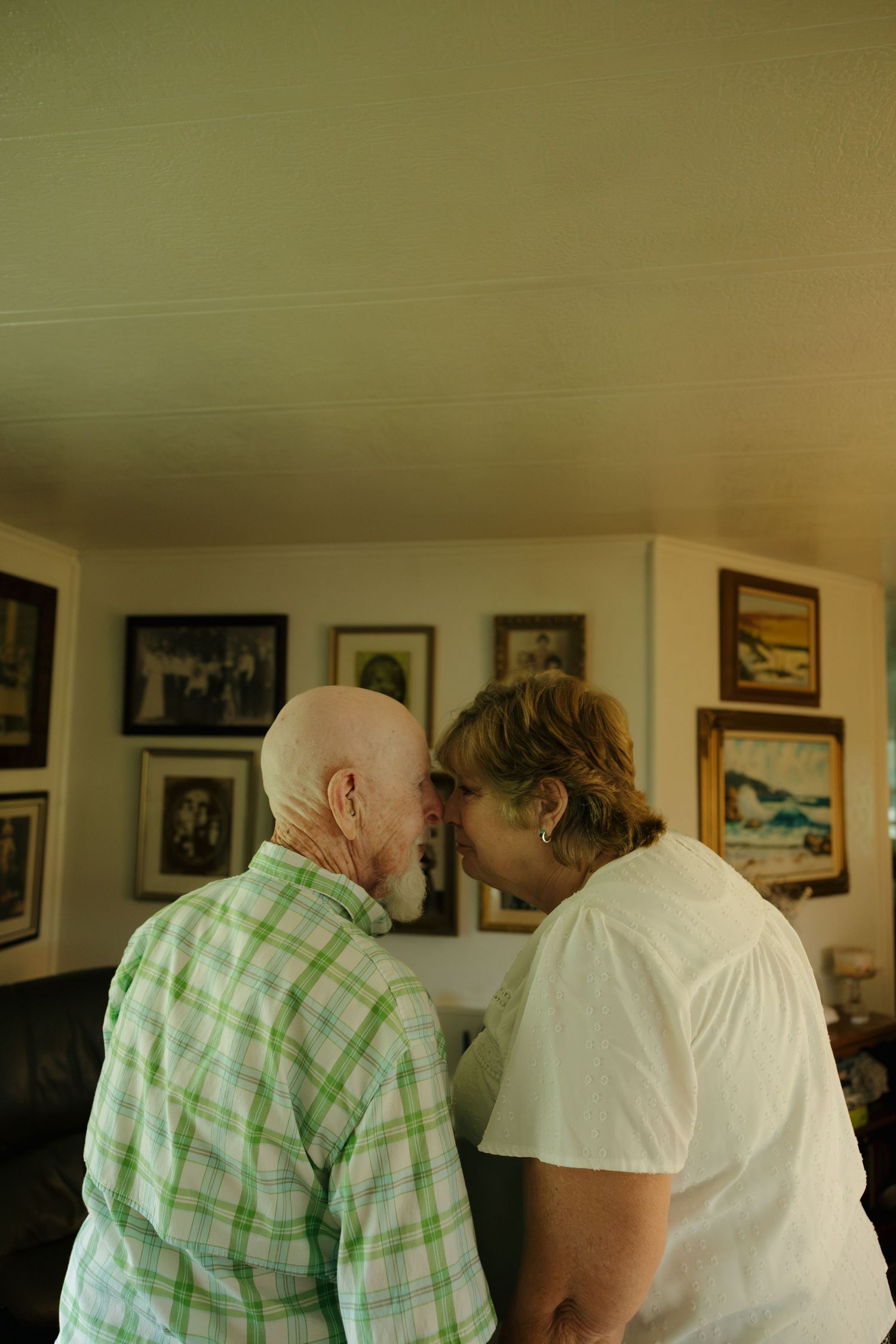 Elderly couple sharing an intimate moment, touching foreheads in a cozy living room with framed artwork on walls.