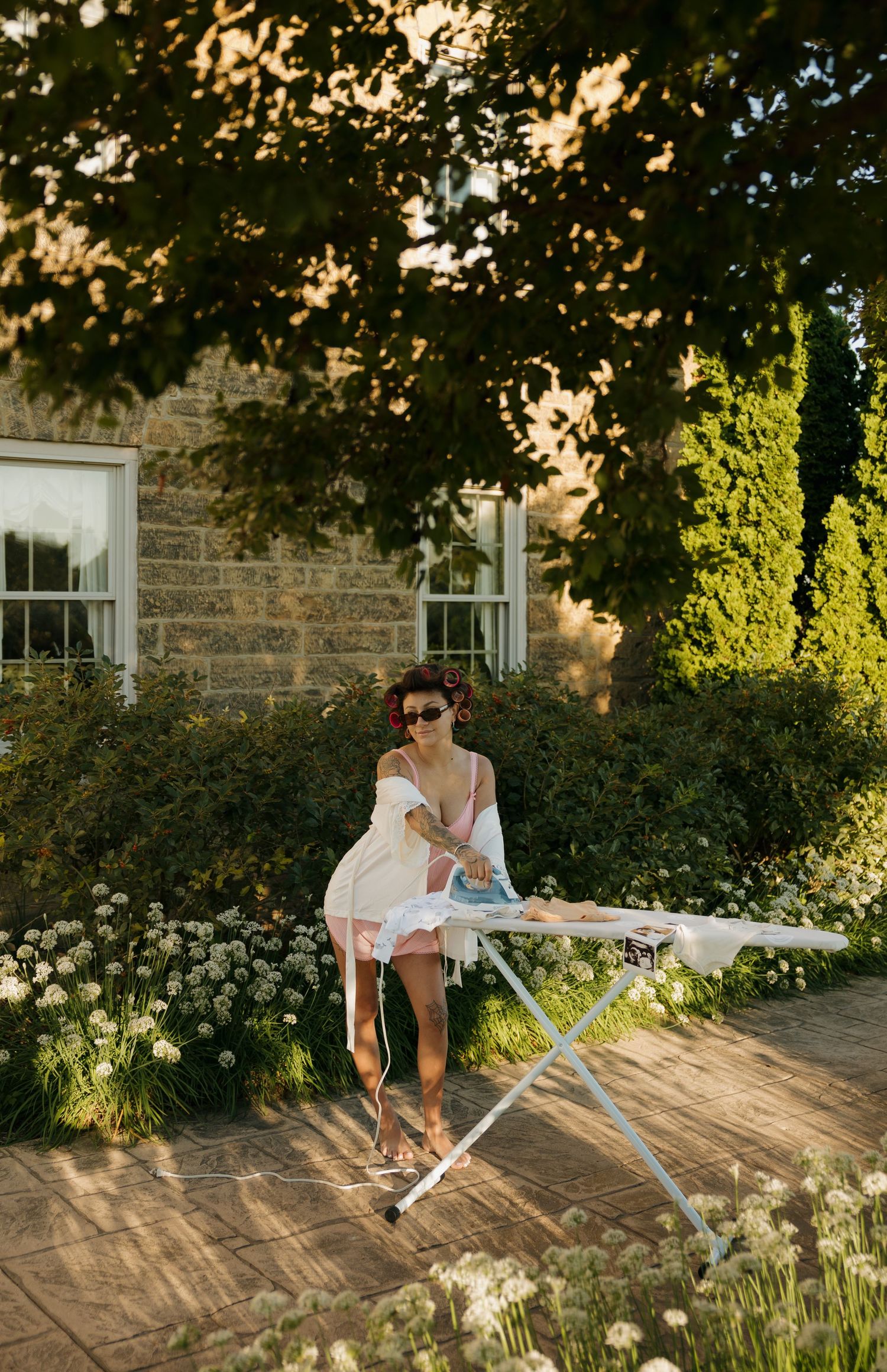 Woman in white dress ironing outdoors in a sunny garden with stone house and lush greenery.