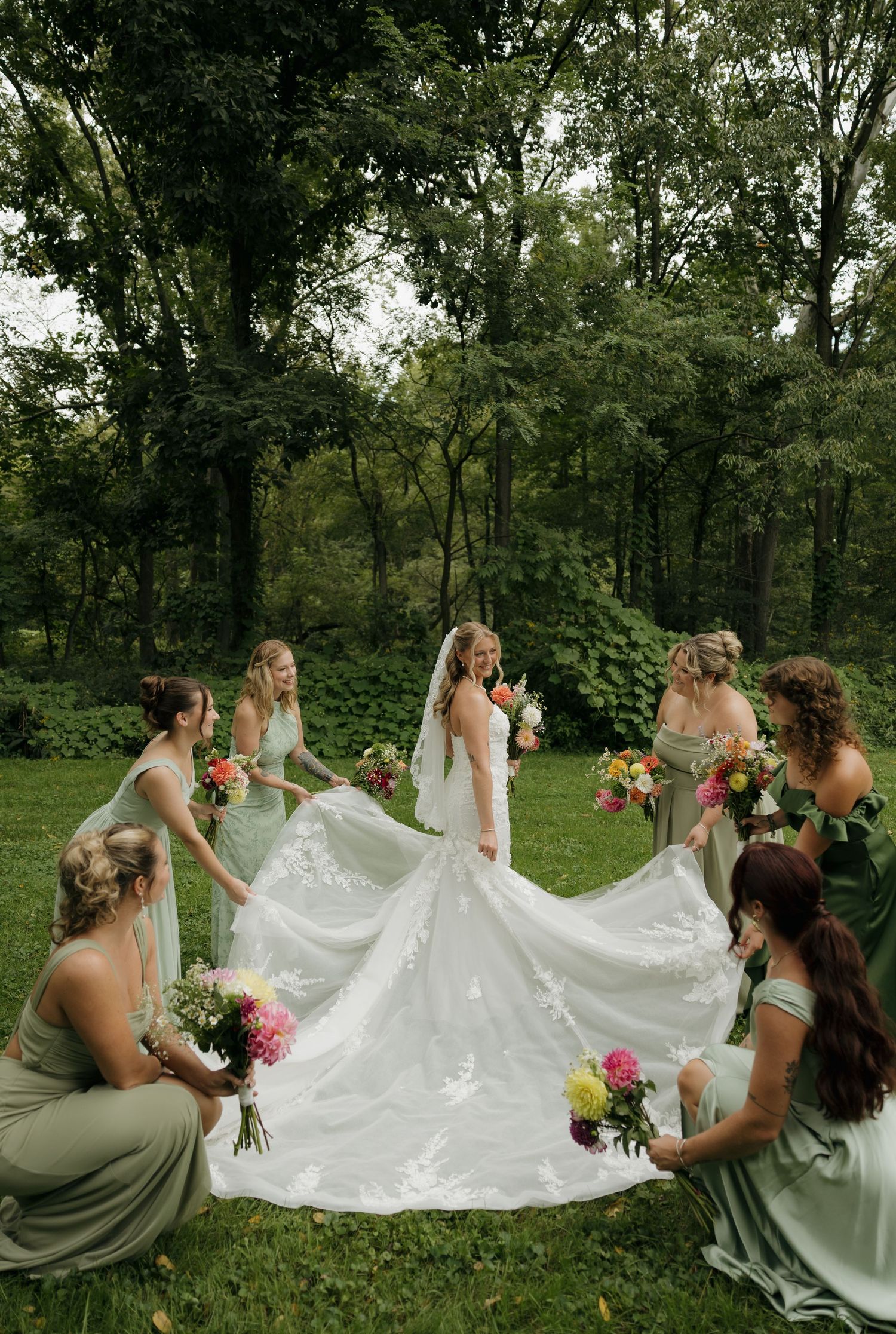 Bride in white gown surrounded by bridesmaids in sage green dresses holding colorful bouquets in a lush garden setting.