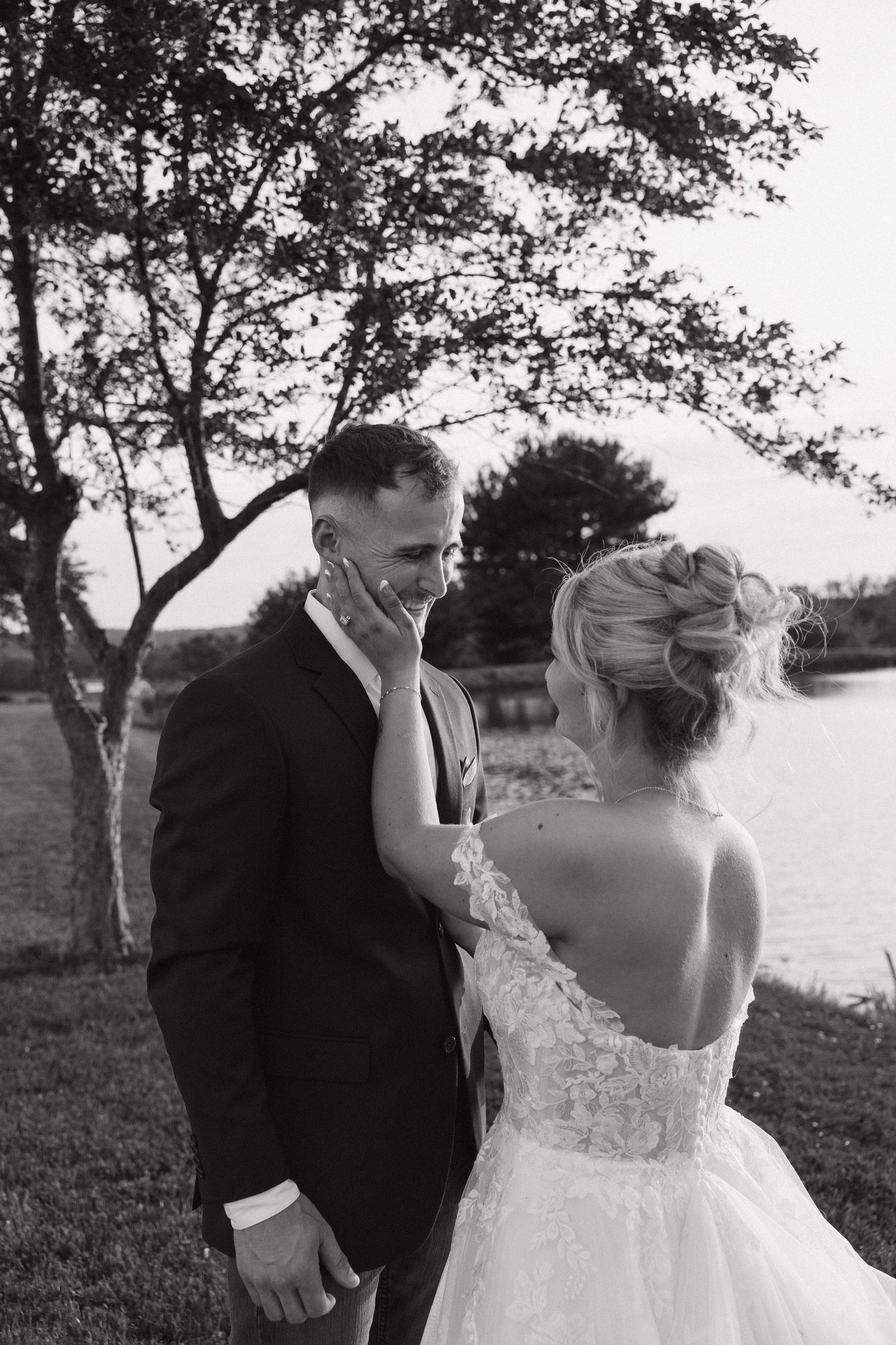 Black and white wedding photo of bride touching grooms face outdoors near a lake with trees in background.
